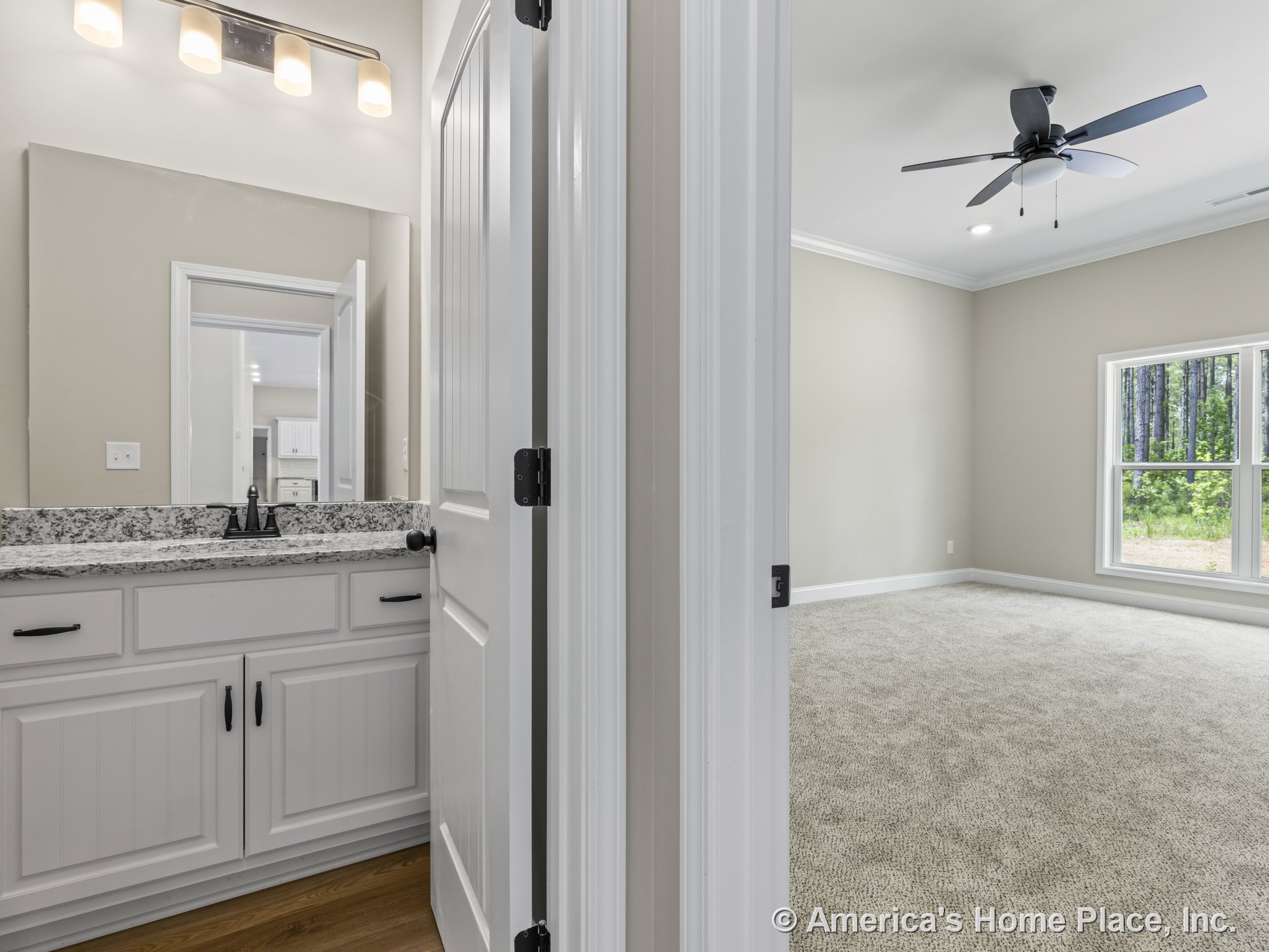 Granite bathroom countertop with white paneled cabinetry and three-light vanity fixture, mirror above sink; adjacent carpeted bedroom featuring large double window with trim