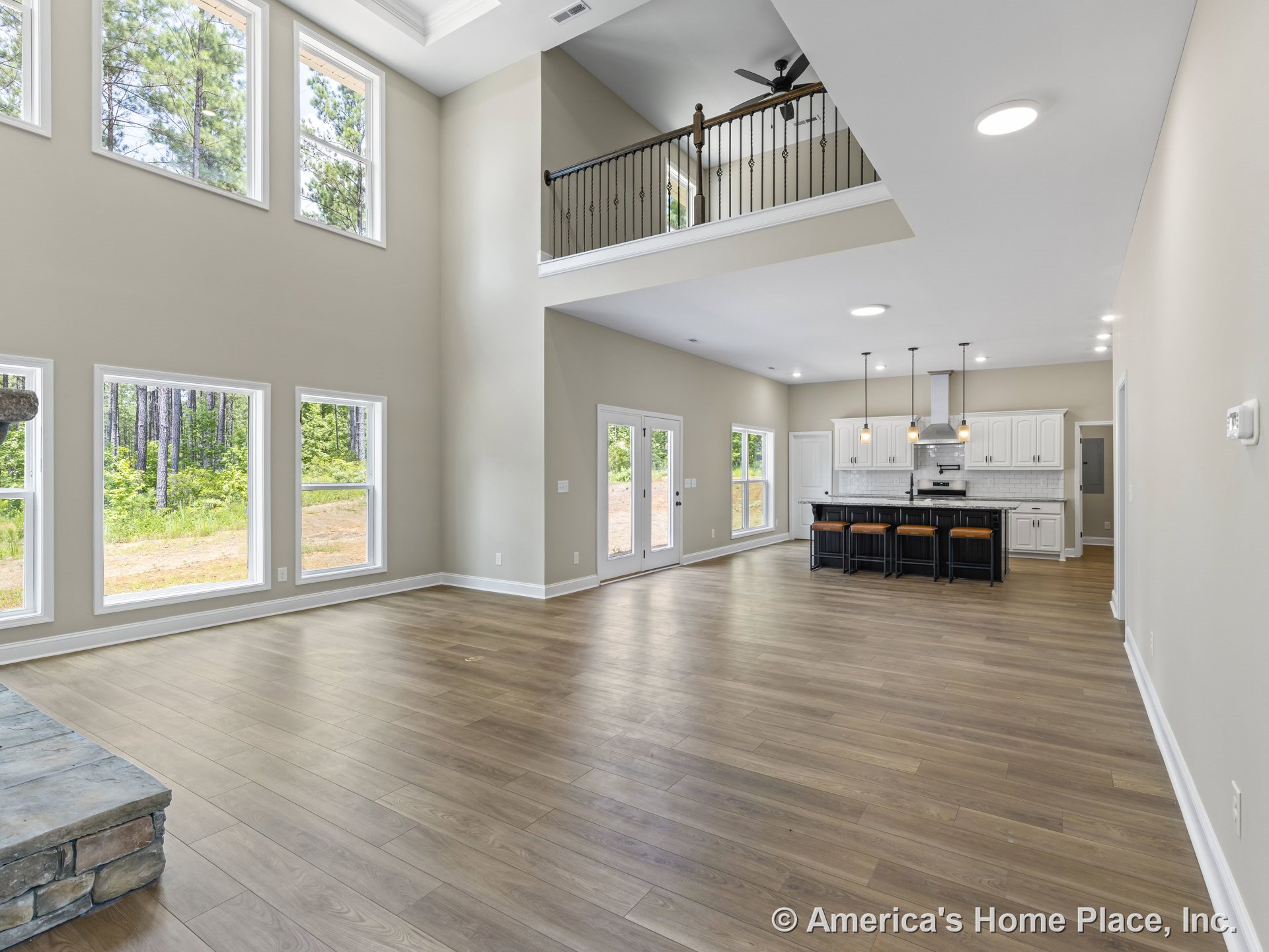 Double-height living area with wall of tall windows, open kitchen featuring white cabinetry and central island, wood-look flooring, stone fireplace hearth, tray ceiling with