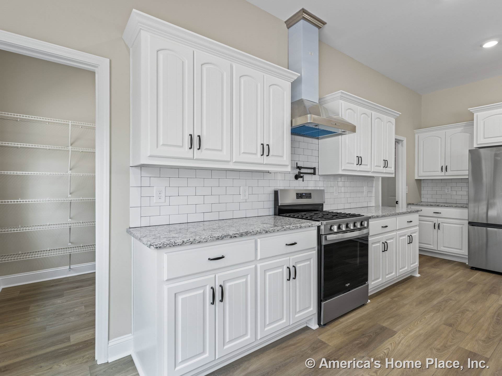 White raised-panel kitchen cabinetry with granite countertops, white subway tile backsplash, stainless steel range and hood, pot filler above stove, walk-in pantry with wire