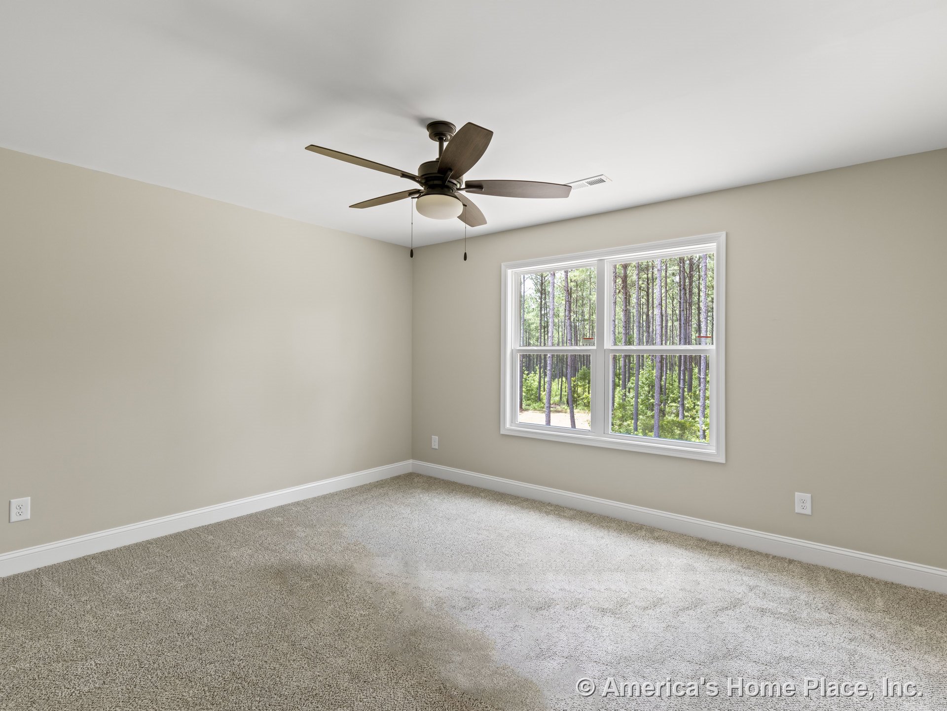 Beige carpeted bedroom with neutral walls, white baseboards, large double window with white trim overlooking wooded scenery, ceiling fan with integrated light fixture, and