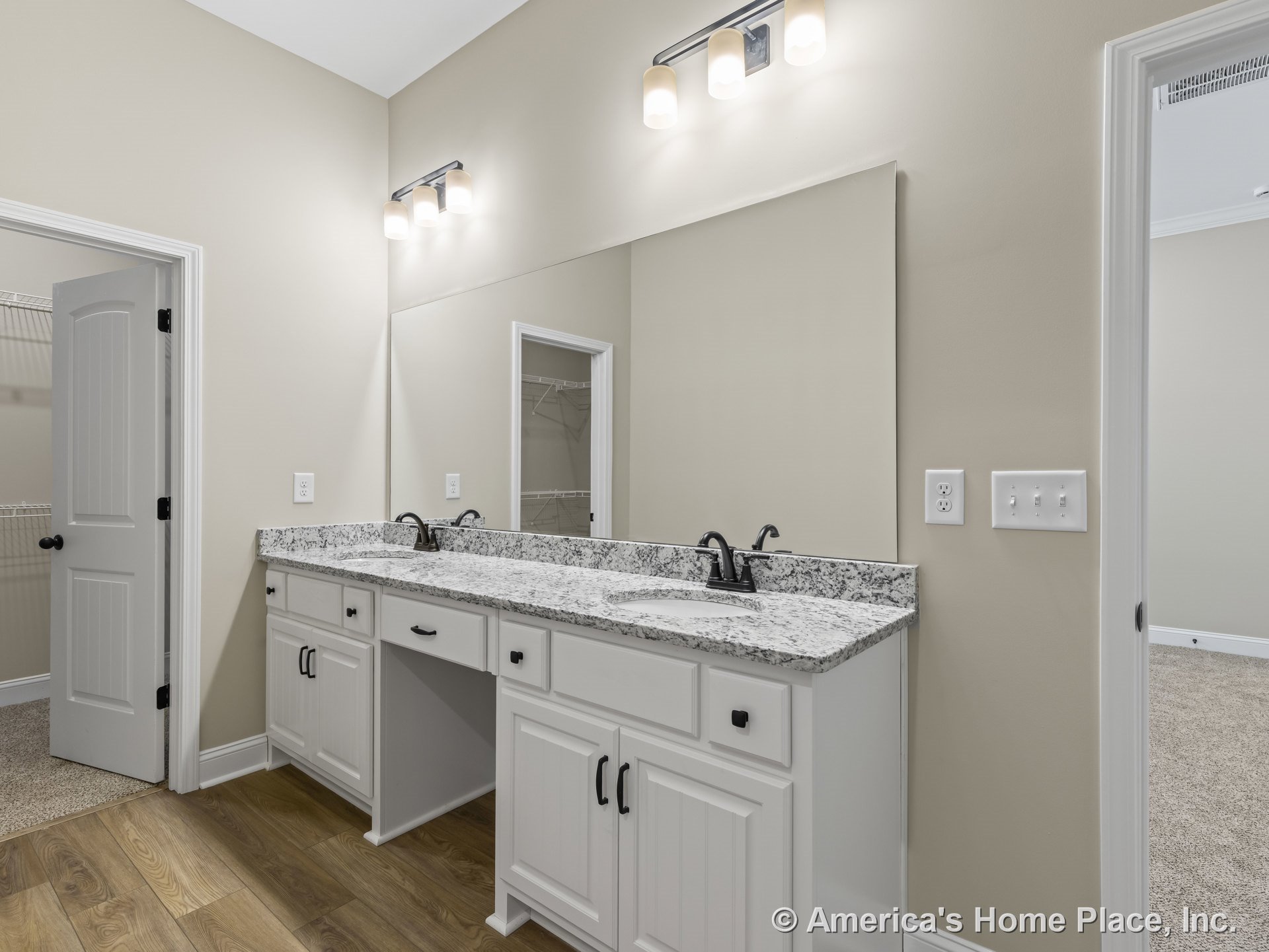 Double vanity with granite countertop, white shaker cabinets, large wall mirror, built-in overhead lighting, wood-look flooring, black faucets, neutral walls, and white trim.