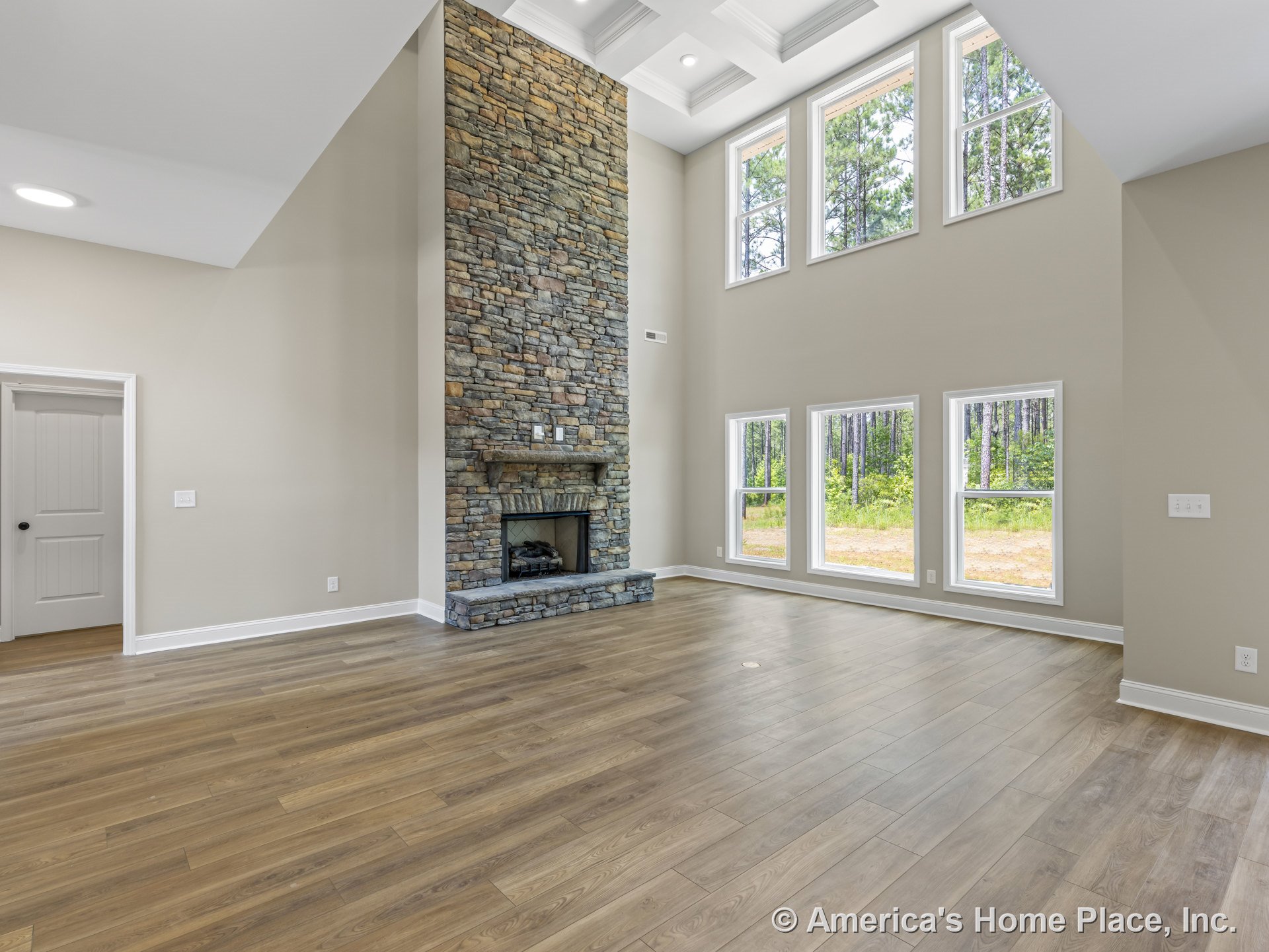 Floor-to-ceiling stone fireplace centered in living room with coffered ceiling, five large windows, wood plank flooring, neutral wall paint, and white trim and baseboards.