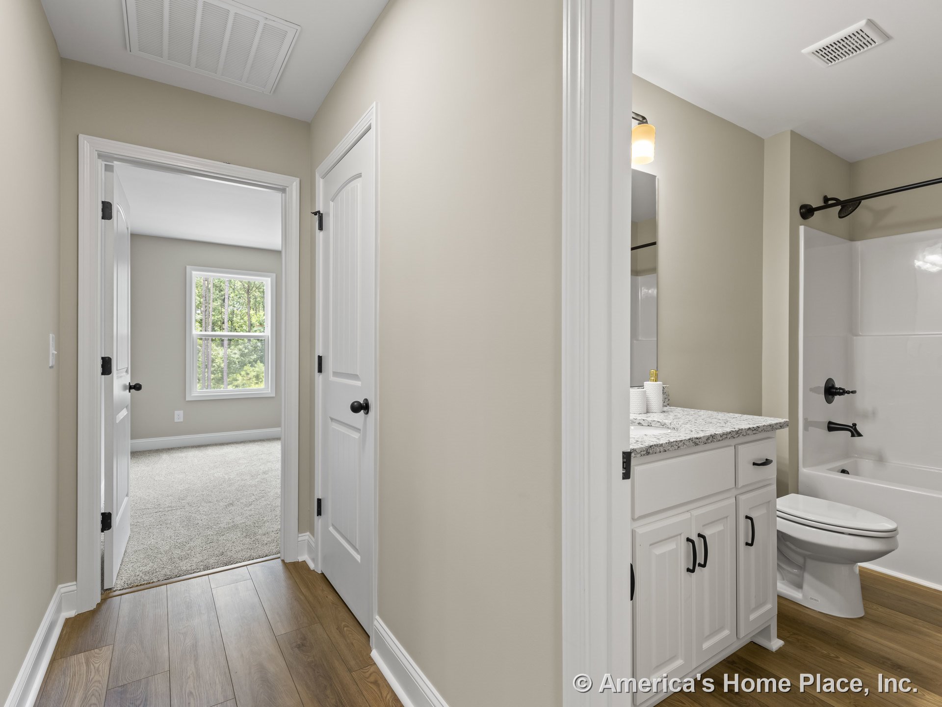 Hallway with wood plank flooring and white paneled doors leading to a bathroom with granite vanity, white cabinetry, bathtub with shower fixtures, and a carpeted bedroom featuring