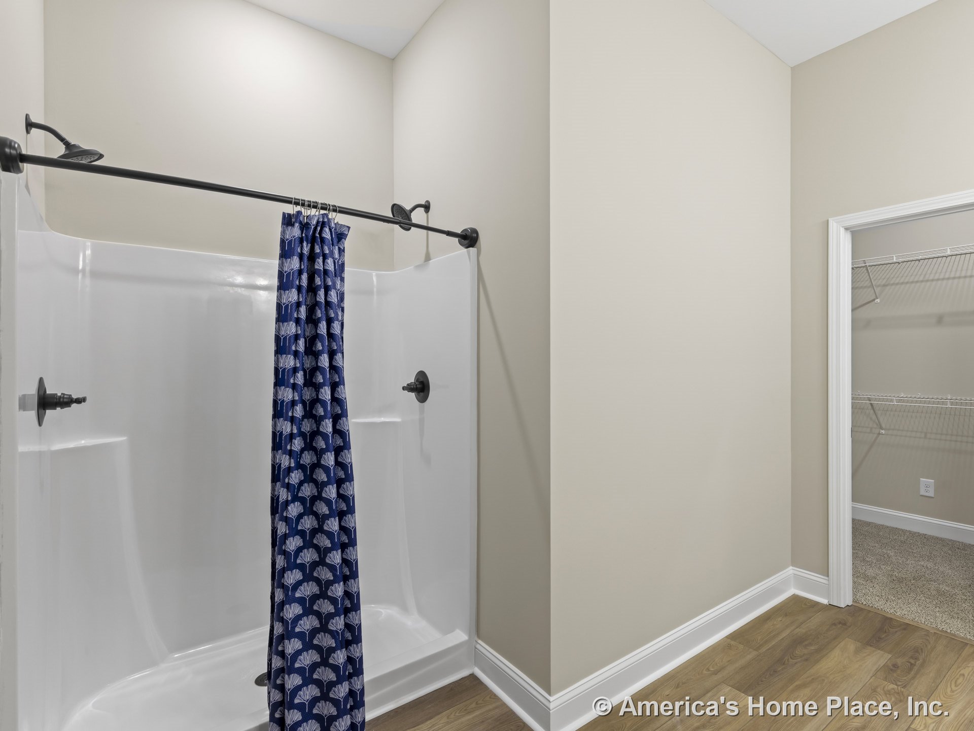 Fiberglass shower enclosure with dual shower heads, adjacent walk-in closet featuring wire shelving, wood-look vinyl flooring, white baseboards, and neutral wall paint.