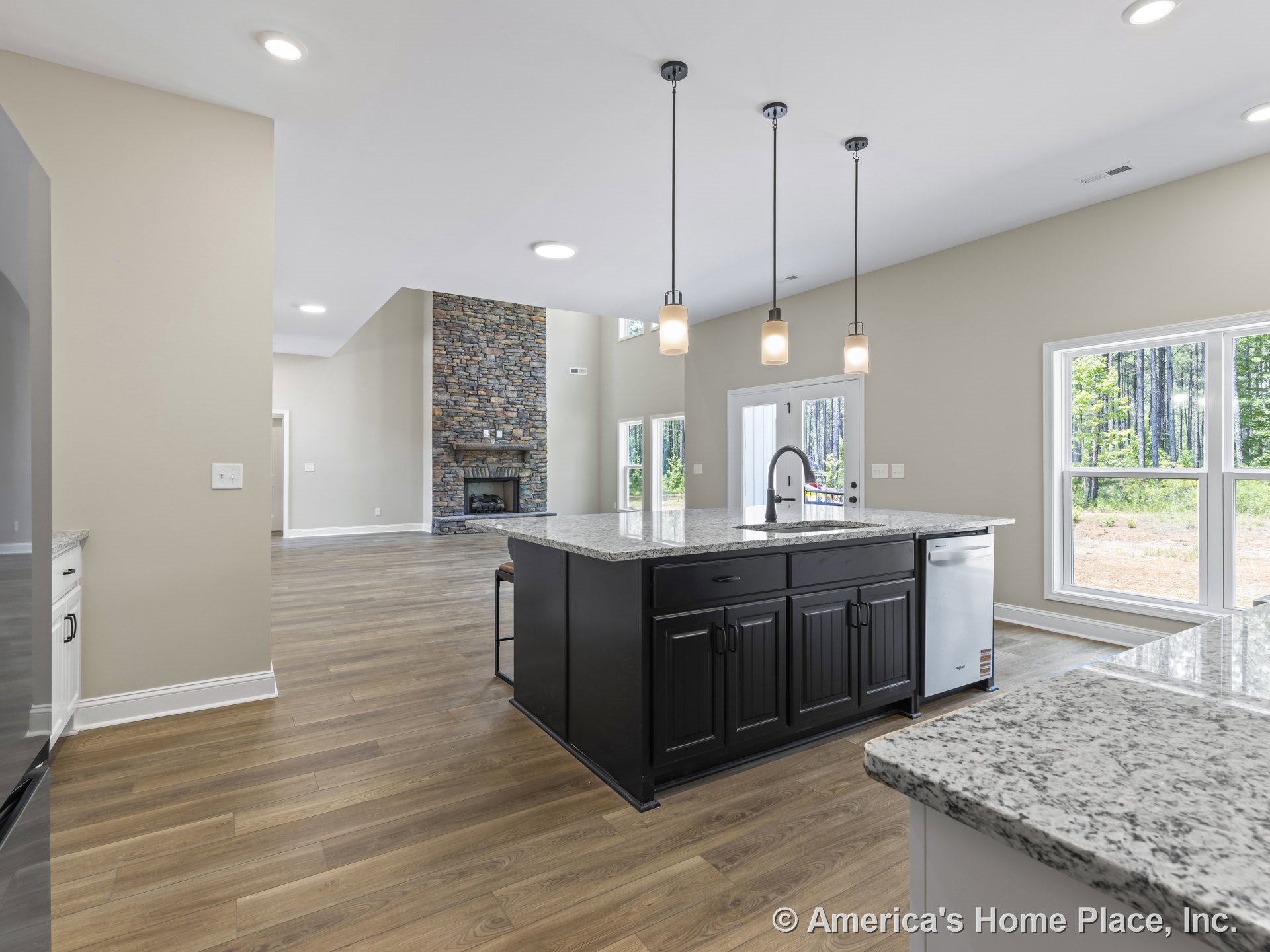 Open kitchen with large granite island featuring black cabinetry and pendant lights, wide plank wood flooring, stone fireplace in adjacent living area, and expansive windows with