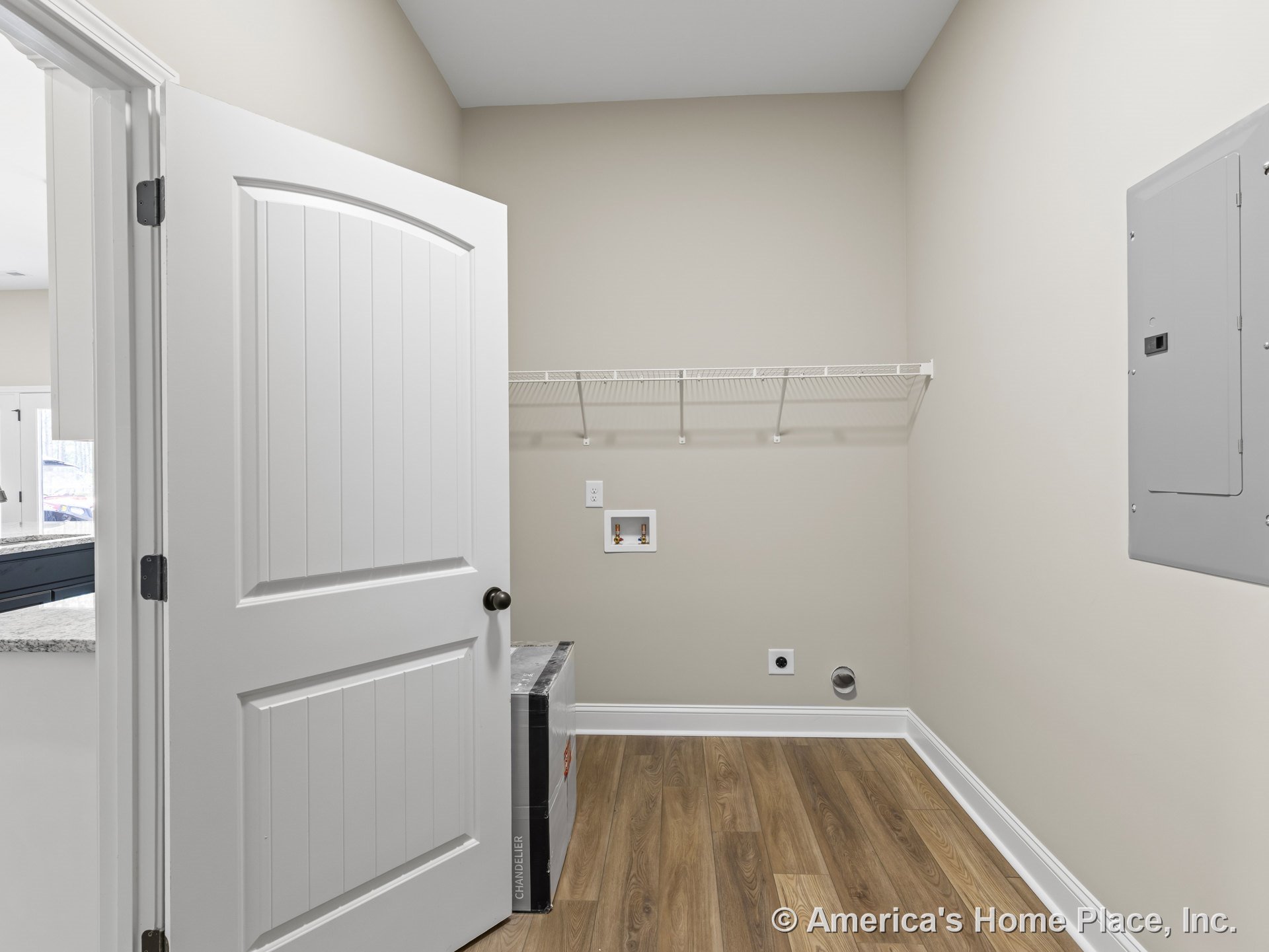 Laundry room with wall-mounted wire shelf, utility hookups, wood-look vinyl flooring, electrical panel, white paneled interior door, neutral painted walls, and white baseboards.