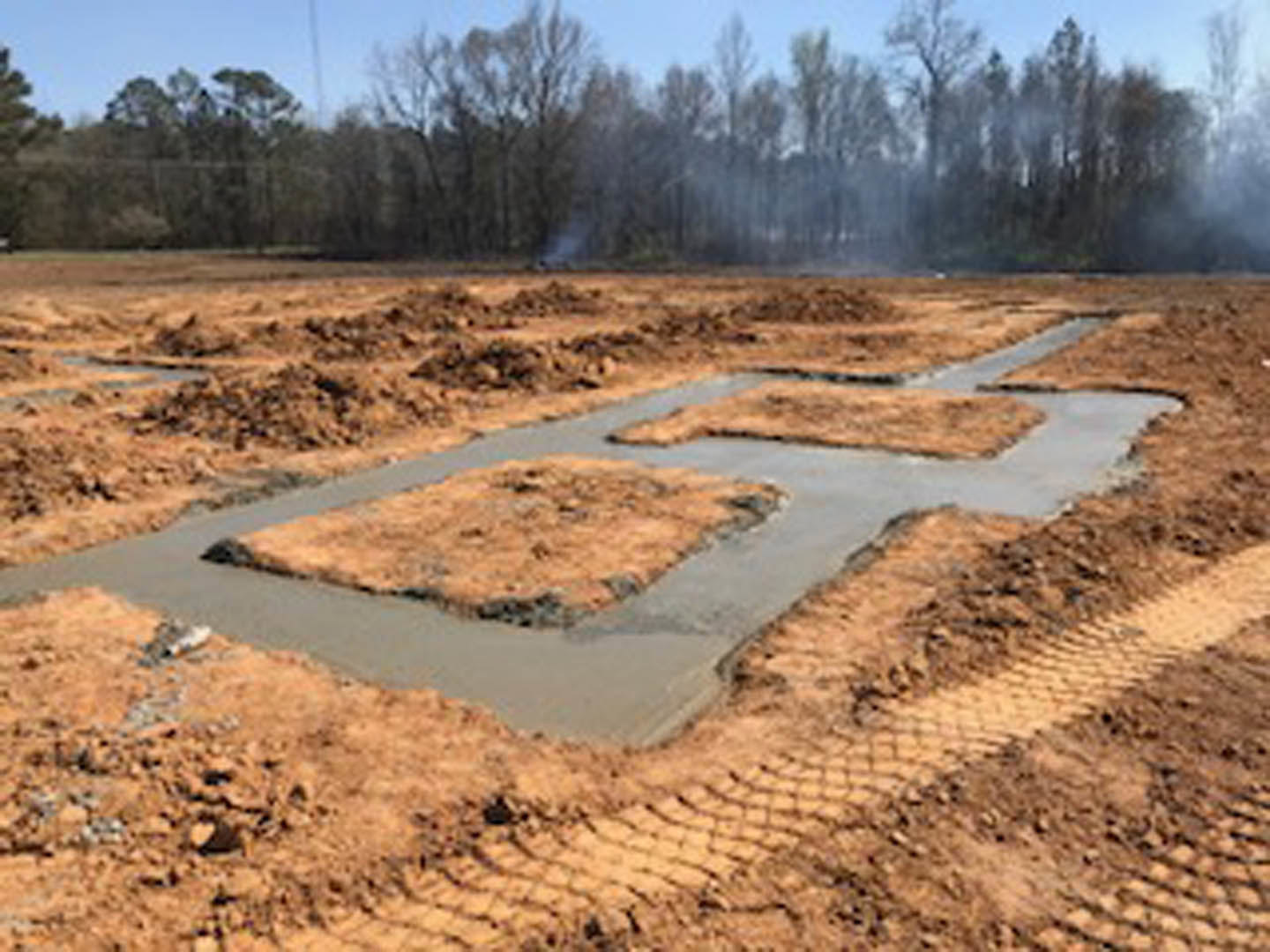 Square concrete foundation surrounded by dirt and tire tracks, with trees and sky in the background