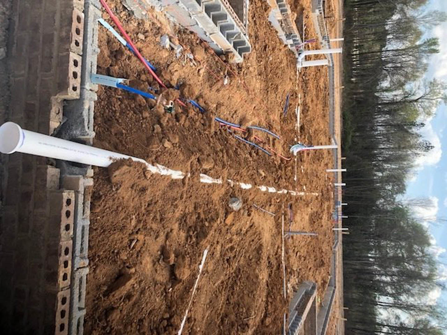 Aerial view of a residential construction site with exposed pipes and electrical wires on bare ground, surrounded by trees.