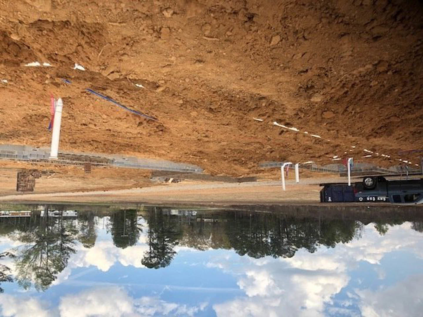 Modern home reflected in calm lake, surrounded by dirt field and trees, pickup truck parked near water, cloudy sky overhead