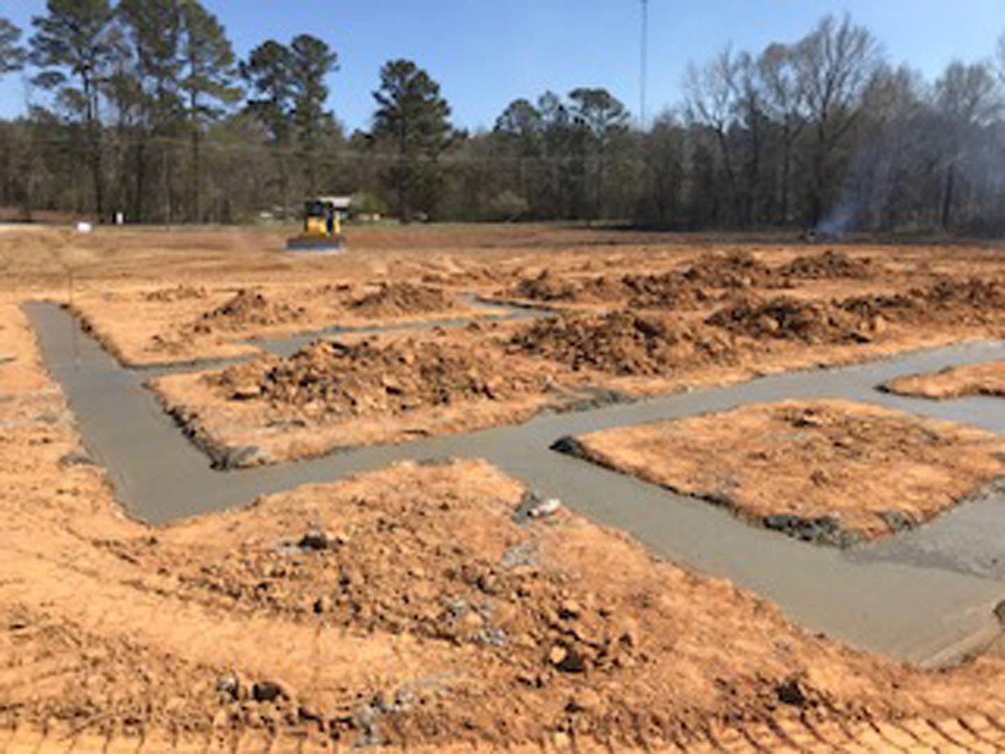 Construction site with exposed soil and mud, shallow ditch in foreground, scattered patches of grass, distant trees under open sky