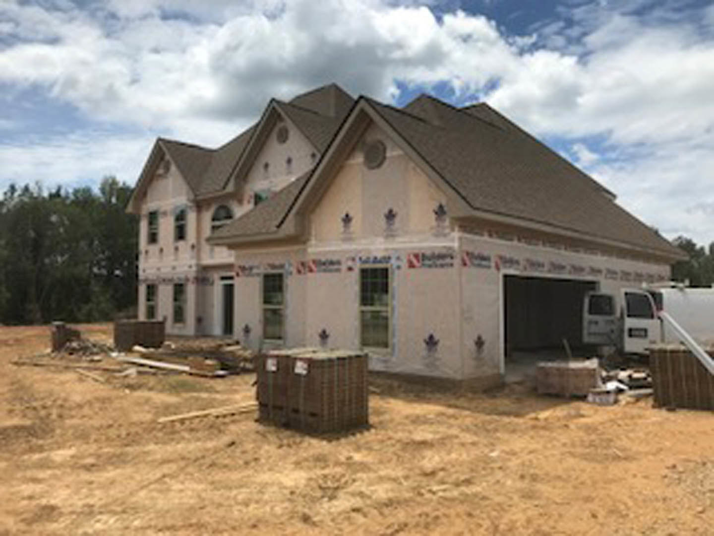 Framed house under construction with exposed wood, attached garage, white van parked on dirt driveway, grassy field and scattered building materials, cloudy sky overhead