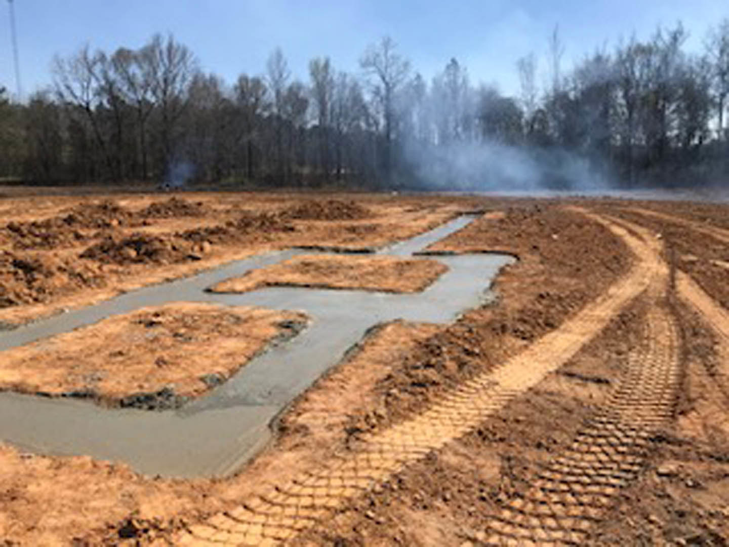 Square concrete foundation slab set in a dirt field, surrounded by muddy ground, tire tracks, and patches of brown grass, with trees and smoke visible in the background.