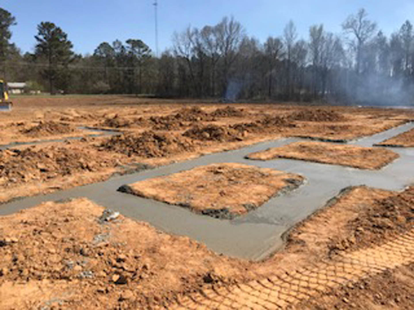 Square dirt field with uneven soil, scattered patches of brown earth, small puddle, and blurred tree in background under open sky
