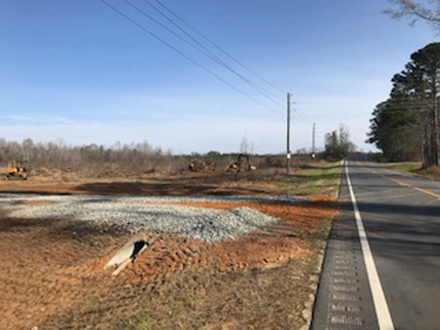 Freshly paved asphalt road bordered by a pile of gravel, power lines overhead, grassy verge and scattered trees under a cloudy sky