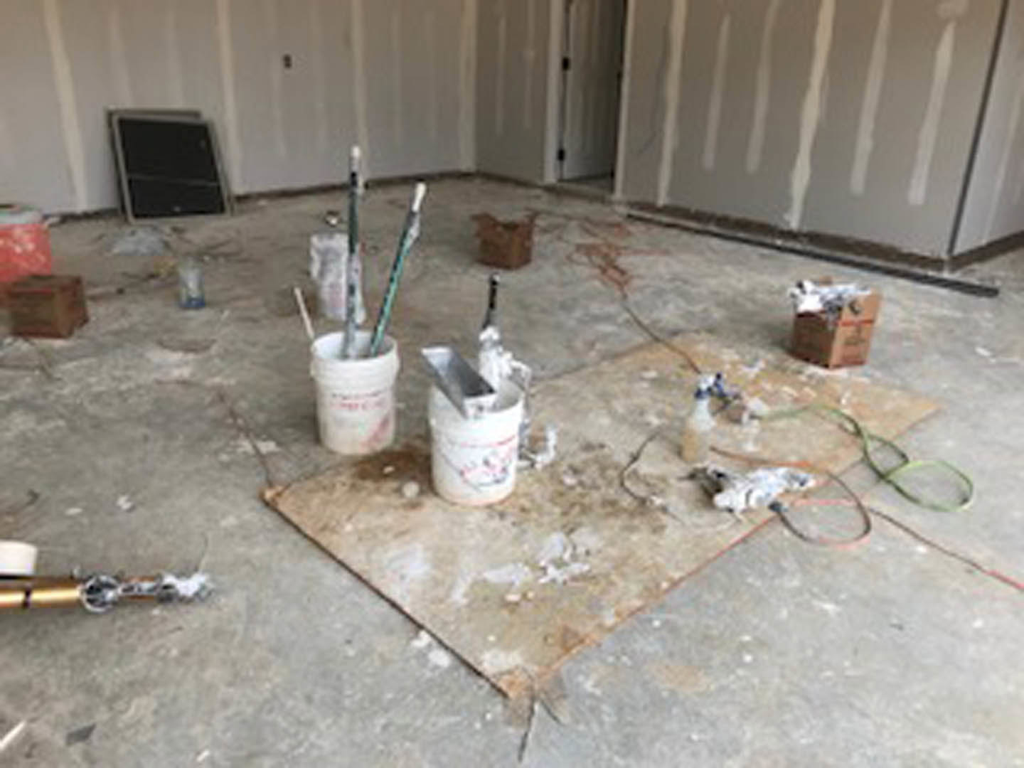 Paint buckets and brushes scattered on a messy floor, white wall partially painted, close-up of white paint can, blurry red and black objects nearby