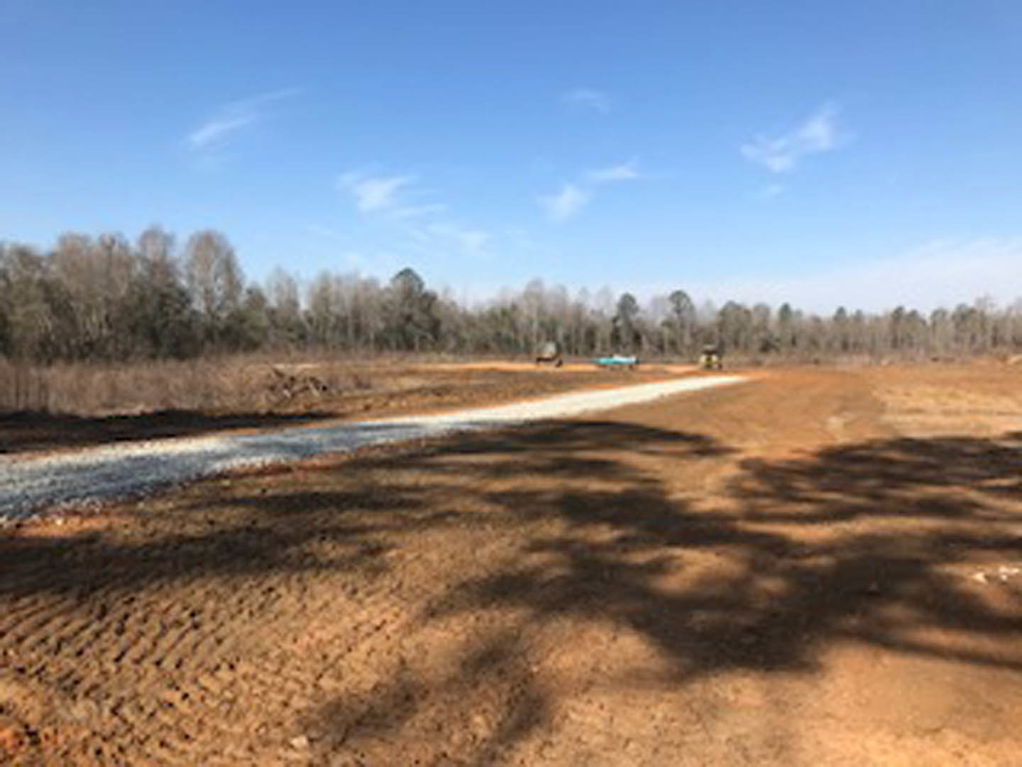 Dirt road bordered by trees under a partly cloudy blue sky, tree shadows cast on the ground, natural landscape with soil and greenery
