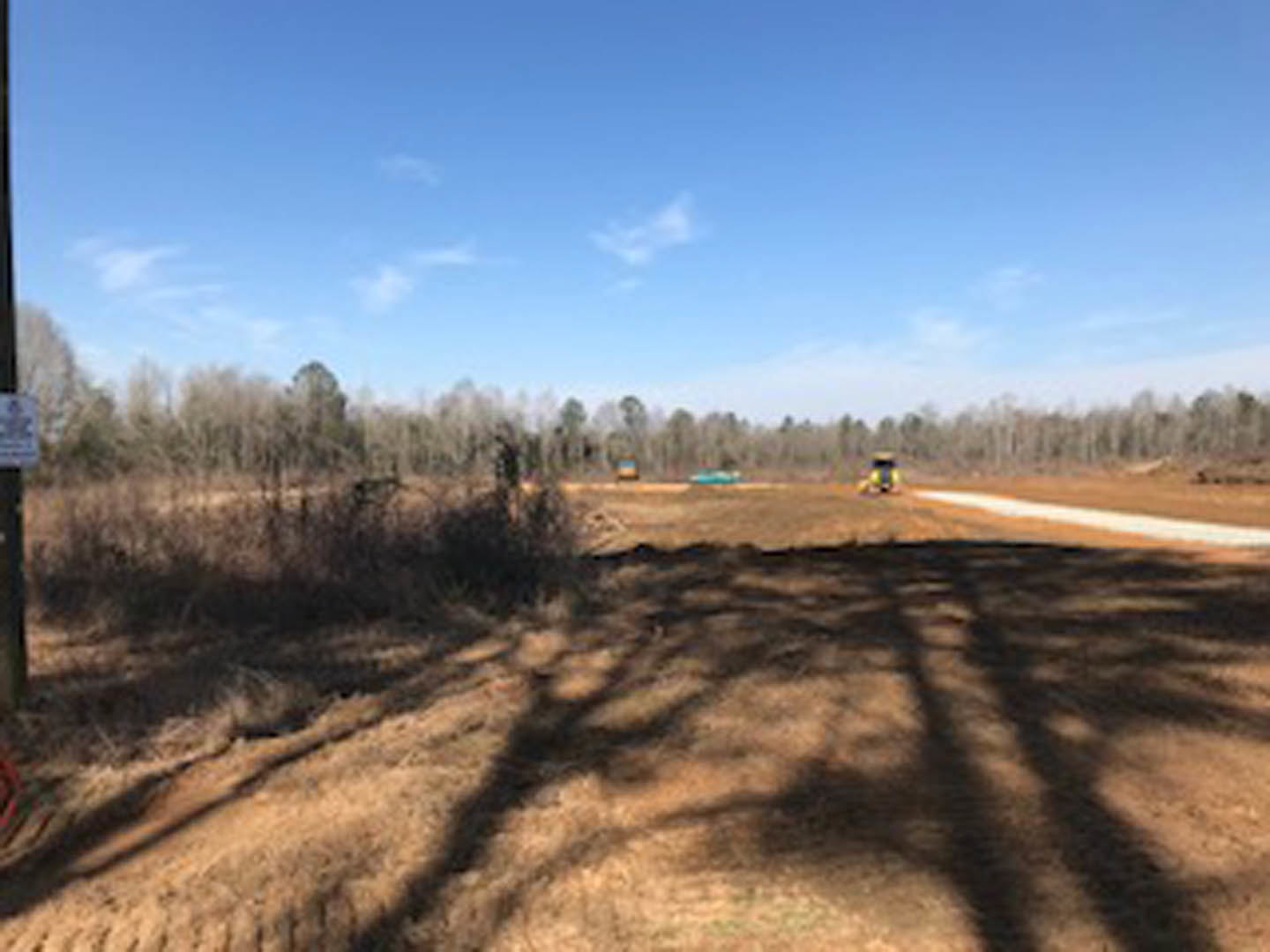 Dirt road bordered by grass and trees under a partly cloudy blue sky, shadow of tree cast on ground, distant building and person’s head blurred in foreground