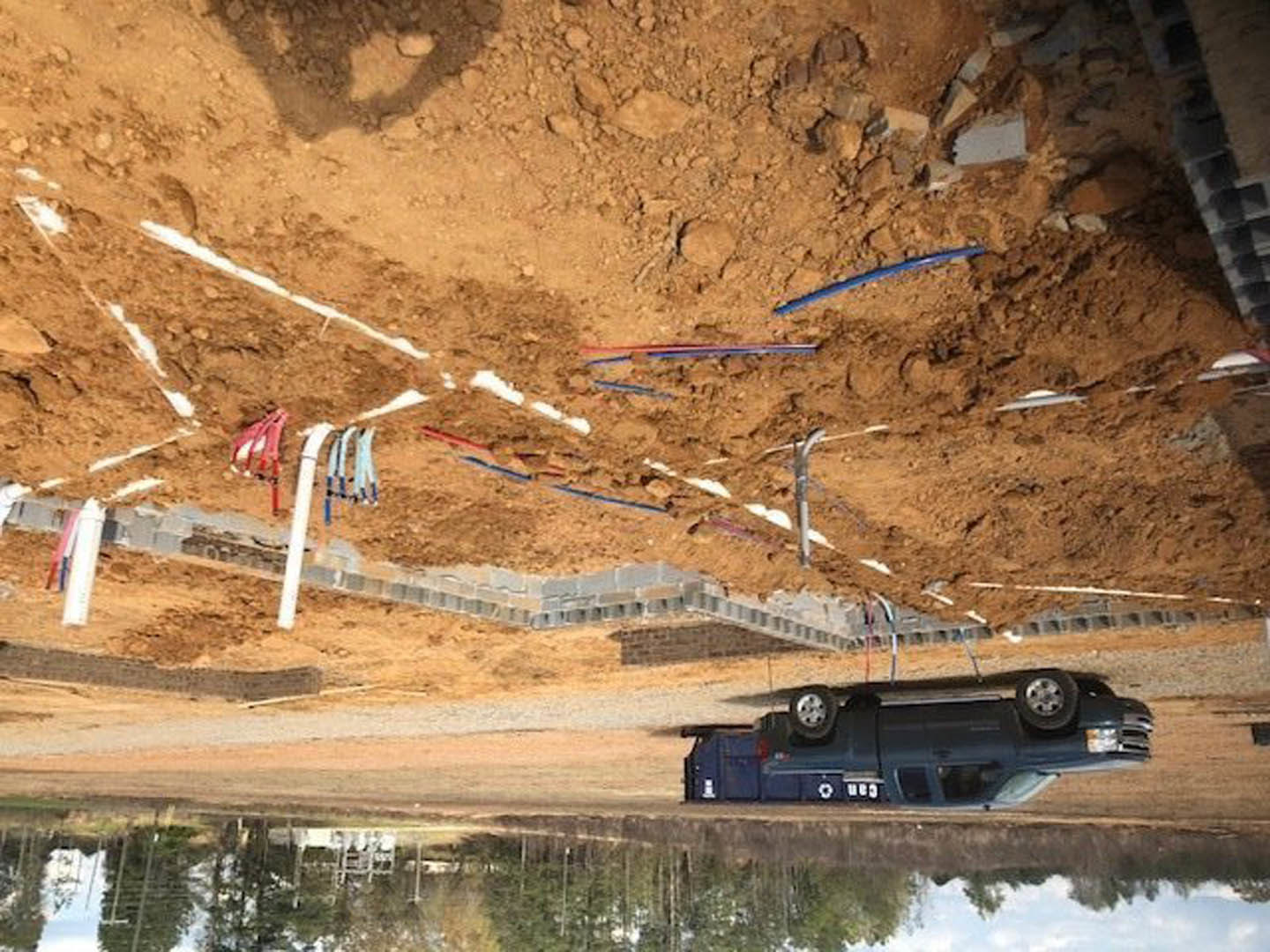 Black pickup truck parked on dirt lot beside blue pipe and white marking line, tire and blue pencil visible in foreground, outdoor construction area.