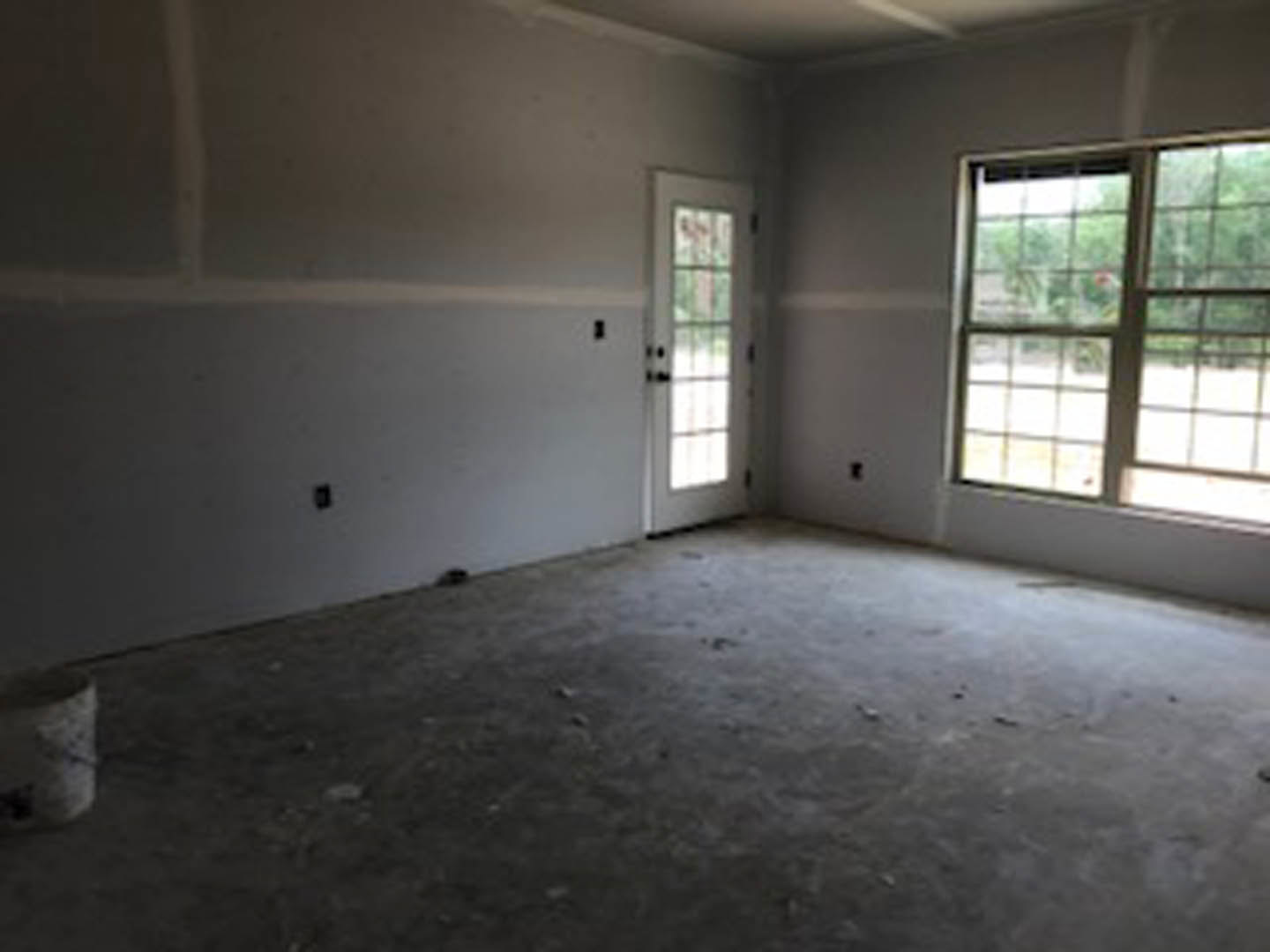 White plaster walls and dirty floor in an empty room with a large window framing leafy trees, a door on the left, and daylight streaming in.