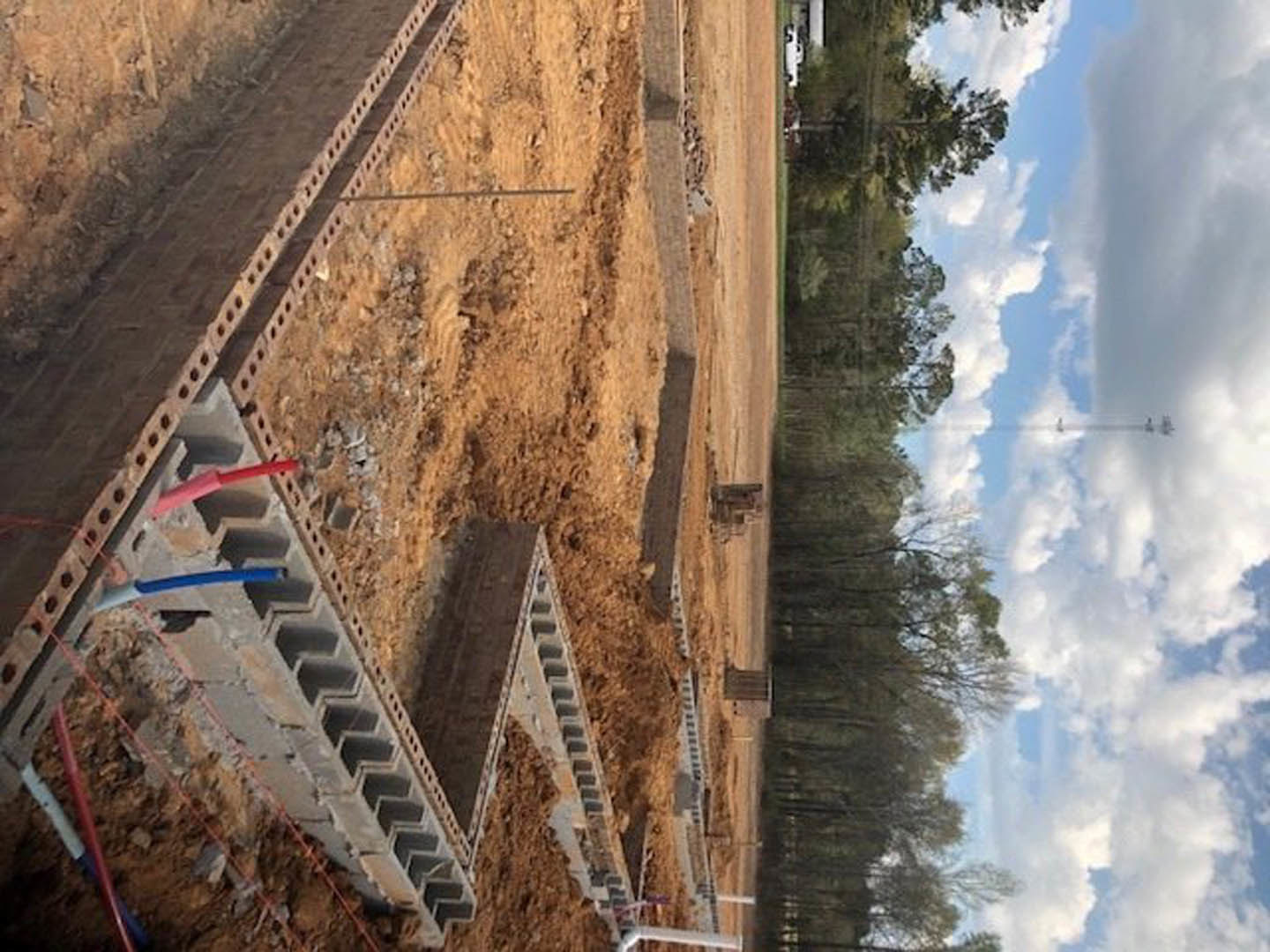Construction site with exposed concrete piles, dirt road, scattered trees, blue sky, and visible power lines overhead