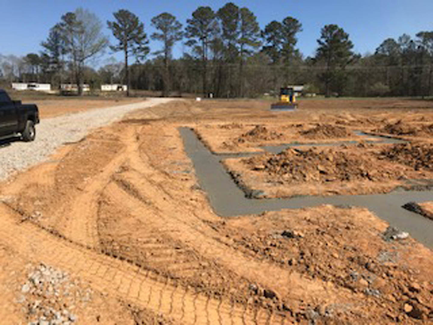 Dirt construction site with a black truck parked near a ditch, yellow vehicle in the background, piles of soil scattered, and a group of trees lining the road.