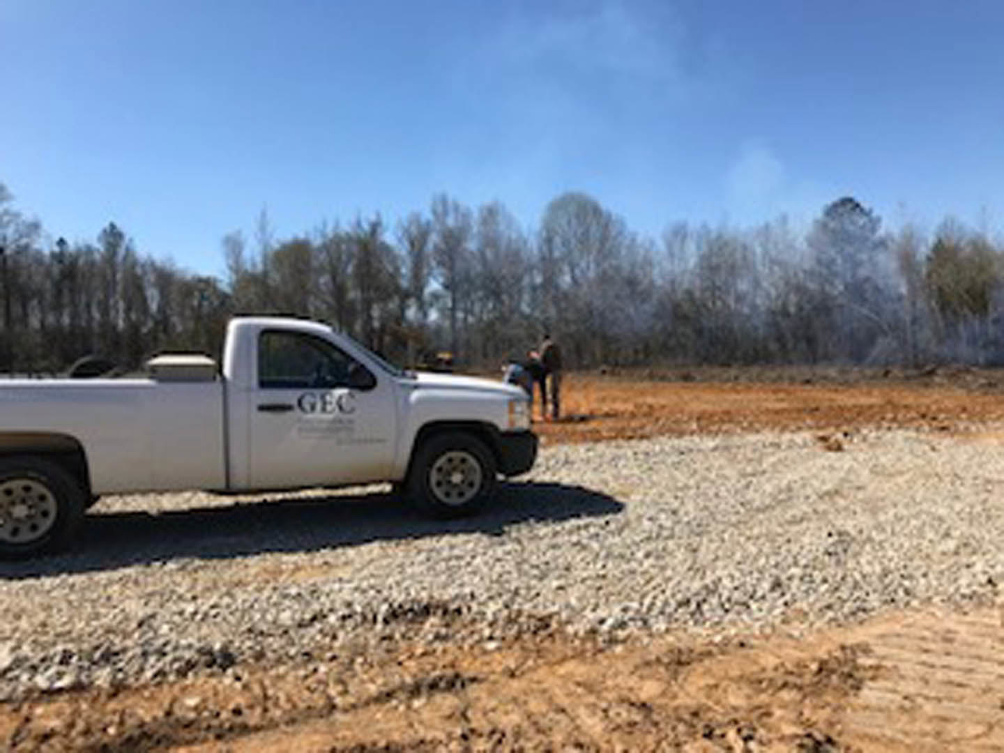 White pickup truck with black lettering parked on a dirt road beside a grove of trees, blurred sign and person visible in the background