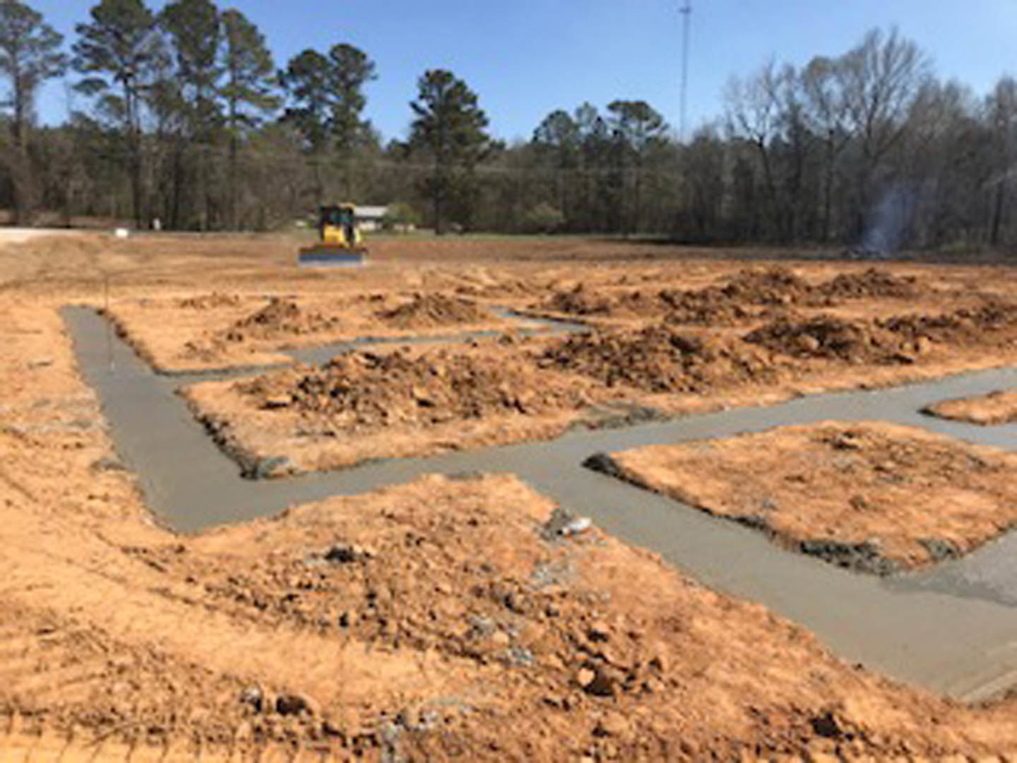 Yellow tractor parked on muddy construction site surrounded by grass, soil, and trees under a cloudy sky