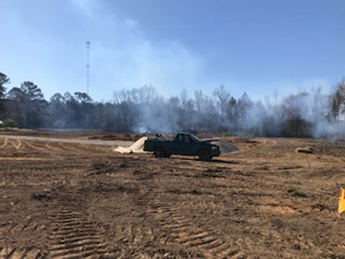 White pickup truck parked on dry grassy field, smoke rising from rear tires, blue sky with scattered clouds, trees in background