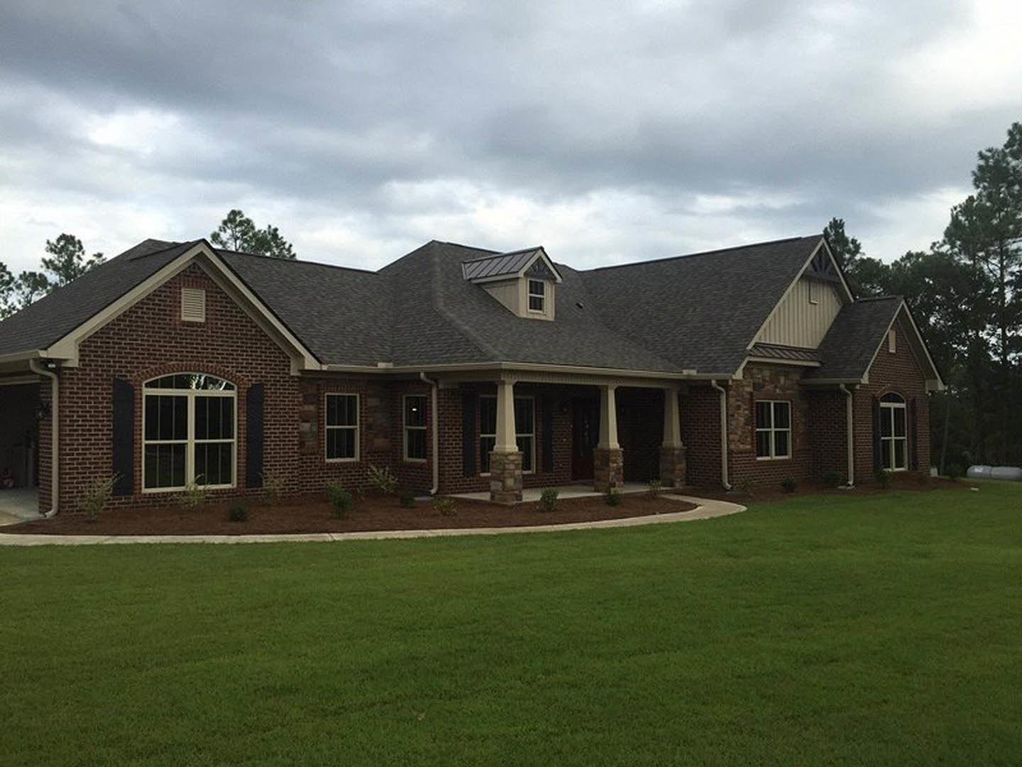 Red brick house with white-framed windows, columned entryway, gabled roof, and manicured green lawn under a cloudy sky