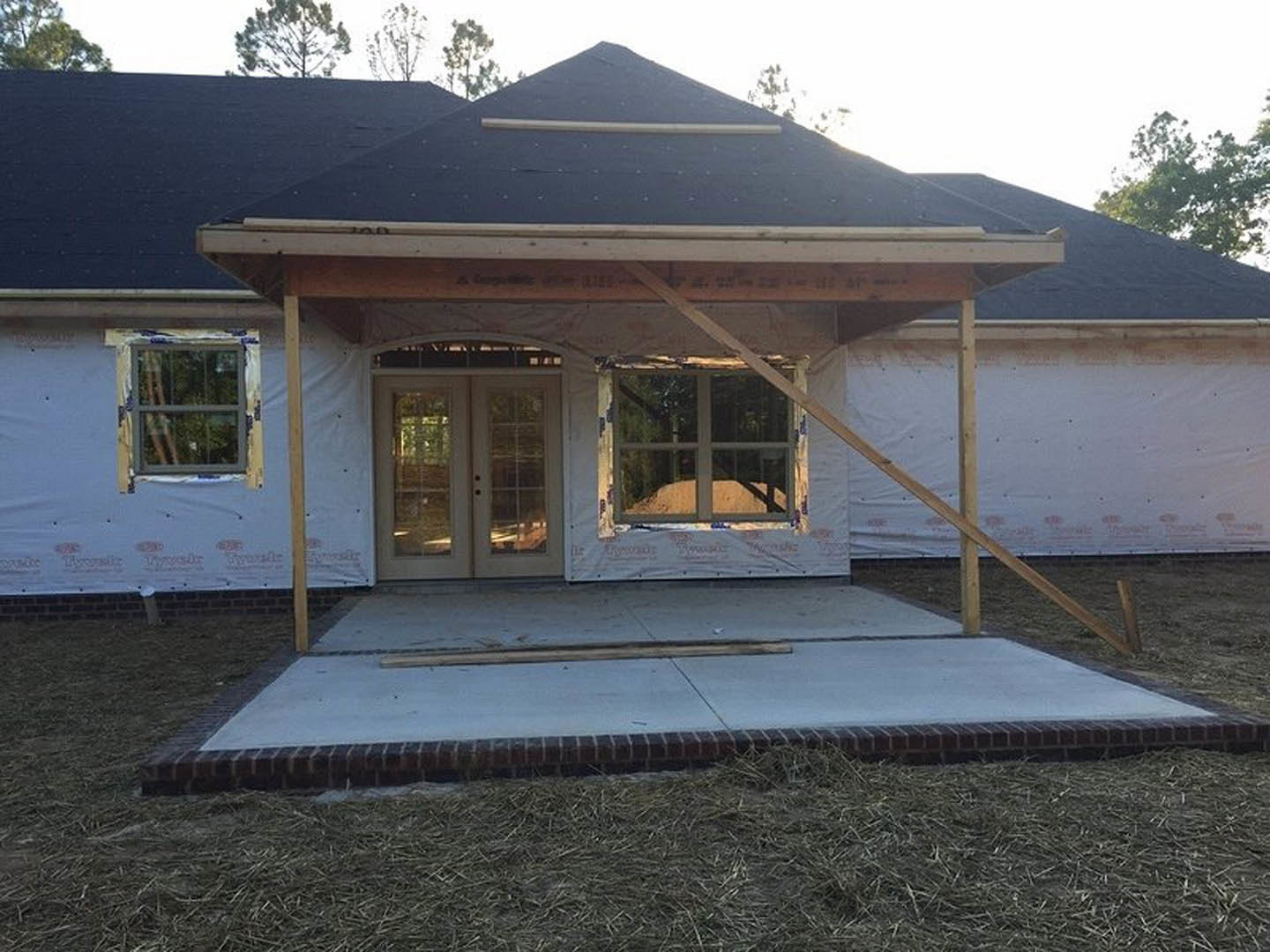 Partially finished home with white exterior wrap, blue-framed window, double glass-paneled doors, concrete sidewalk bordered by grass, and visible roof structure