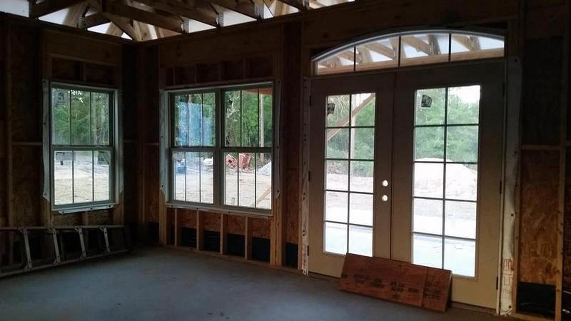 Spacious room with light wood floors, white walls, large windows showing leafy trees, and double glass-paneled doors