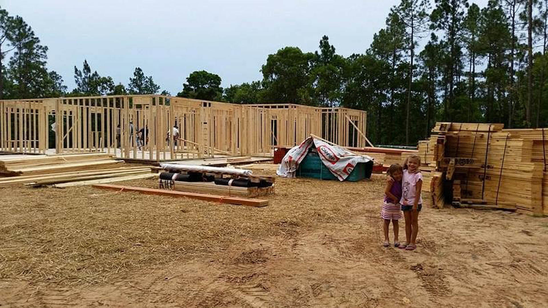 Two girls standing together in front of a partially framed wooden custom home with exposed lumber, construction materials, and a white and red sign on a rope; trees and sky visible