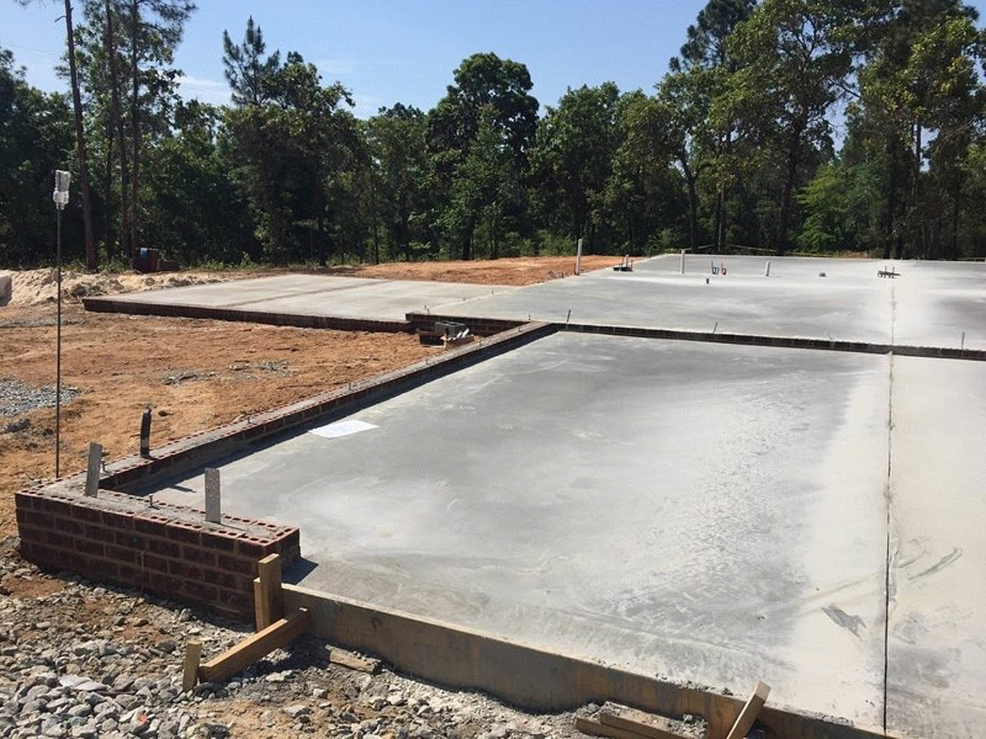 Concrete foundation slab under construction with exposed rebar, surrounded by dirt and trees, adjacent to a brick wall with metal posts.