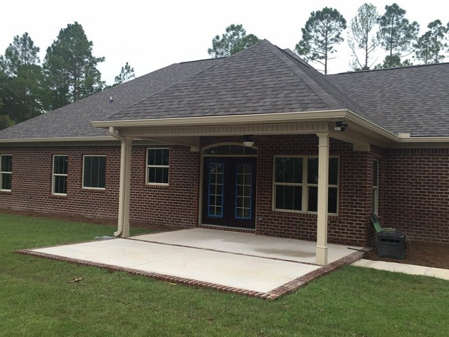 Red brick house with white-framed windows, covered porch, concrete driveway bordered by grass and brick, green chair beside black trash can, shingled roof, leafy tree in yard