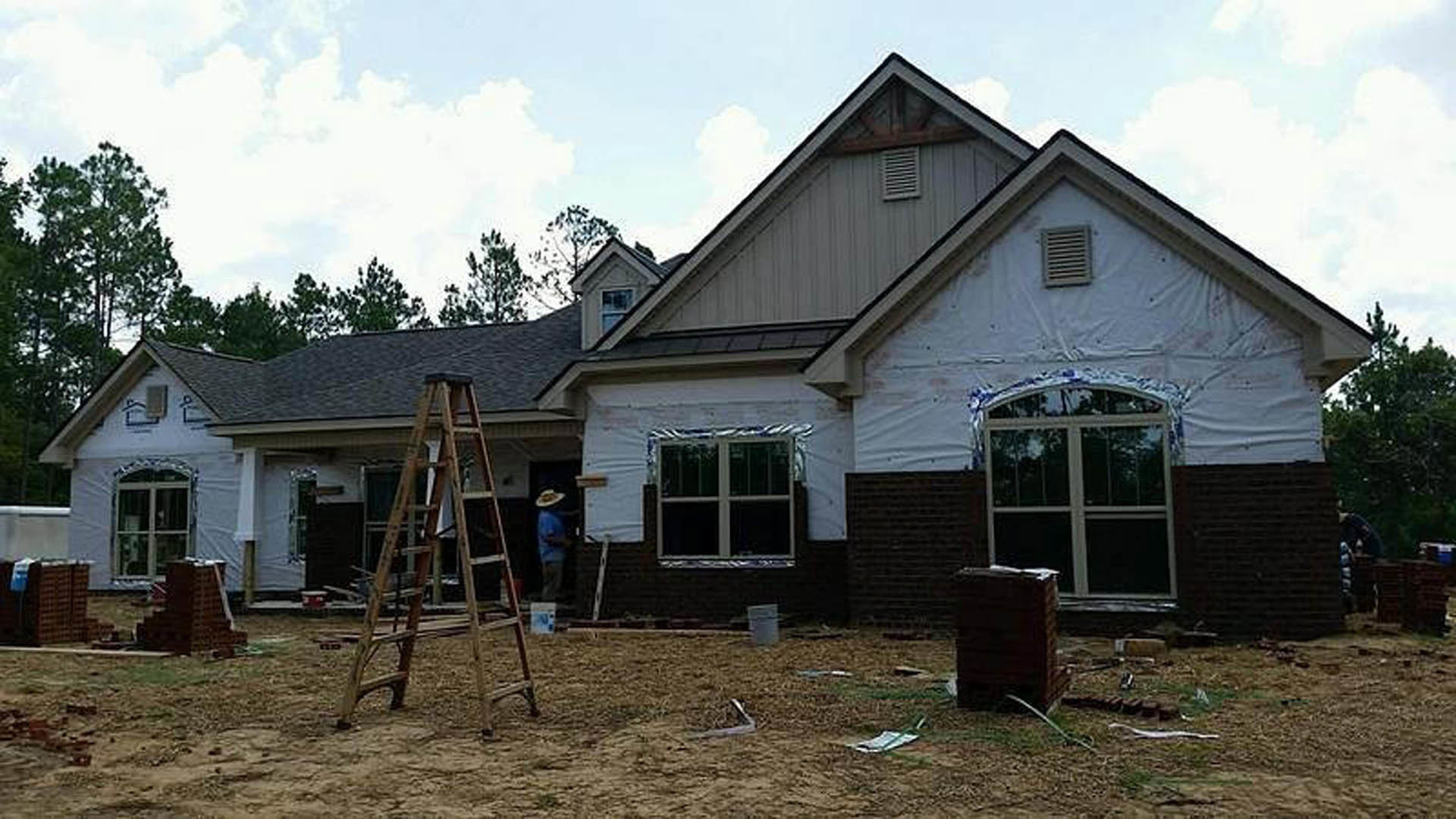 Partially finished exterior with white-framed window, wall vent, ladder leaning against siding, grey barrel in foreground, trees and cloudy sky in background