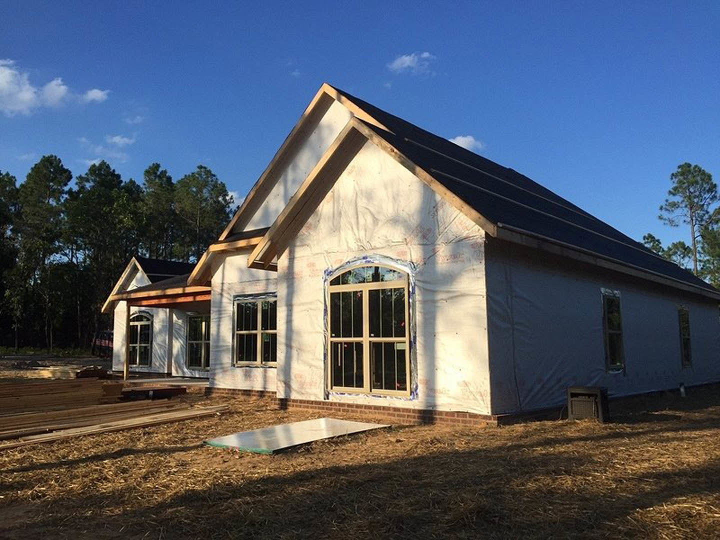 Framed custom home under construction with exposed wood beams, unfinished exterior walls, and surrounding mature trees against a blue sky