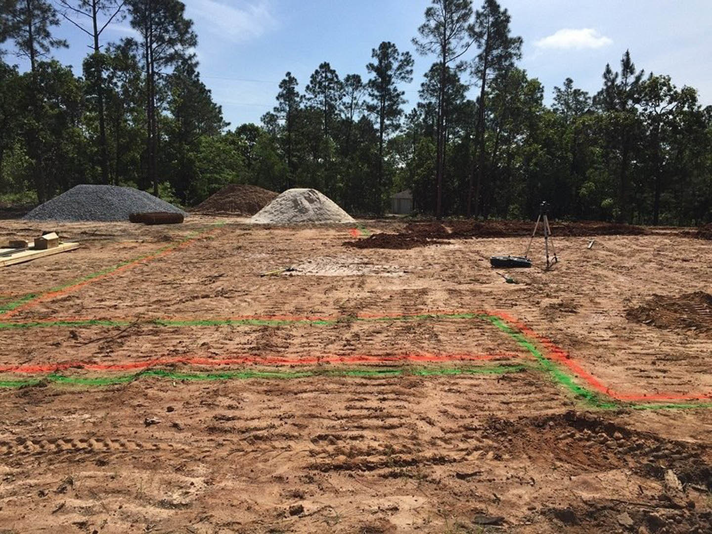 Dirt field with scattered piles of soil, mature trees in the background, and a small building set among grassy land under a partly cloudy sky