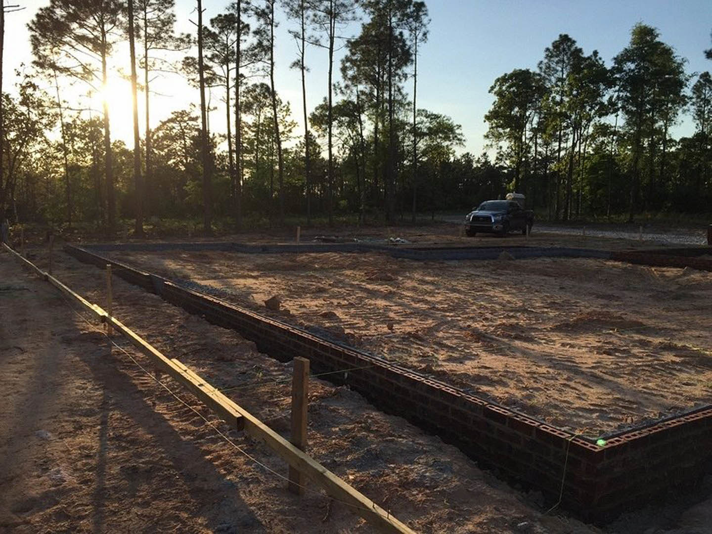 Black car parked on dirt near construction site with metal fence, train tracks, and cluster of trees under clear sky