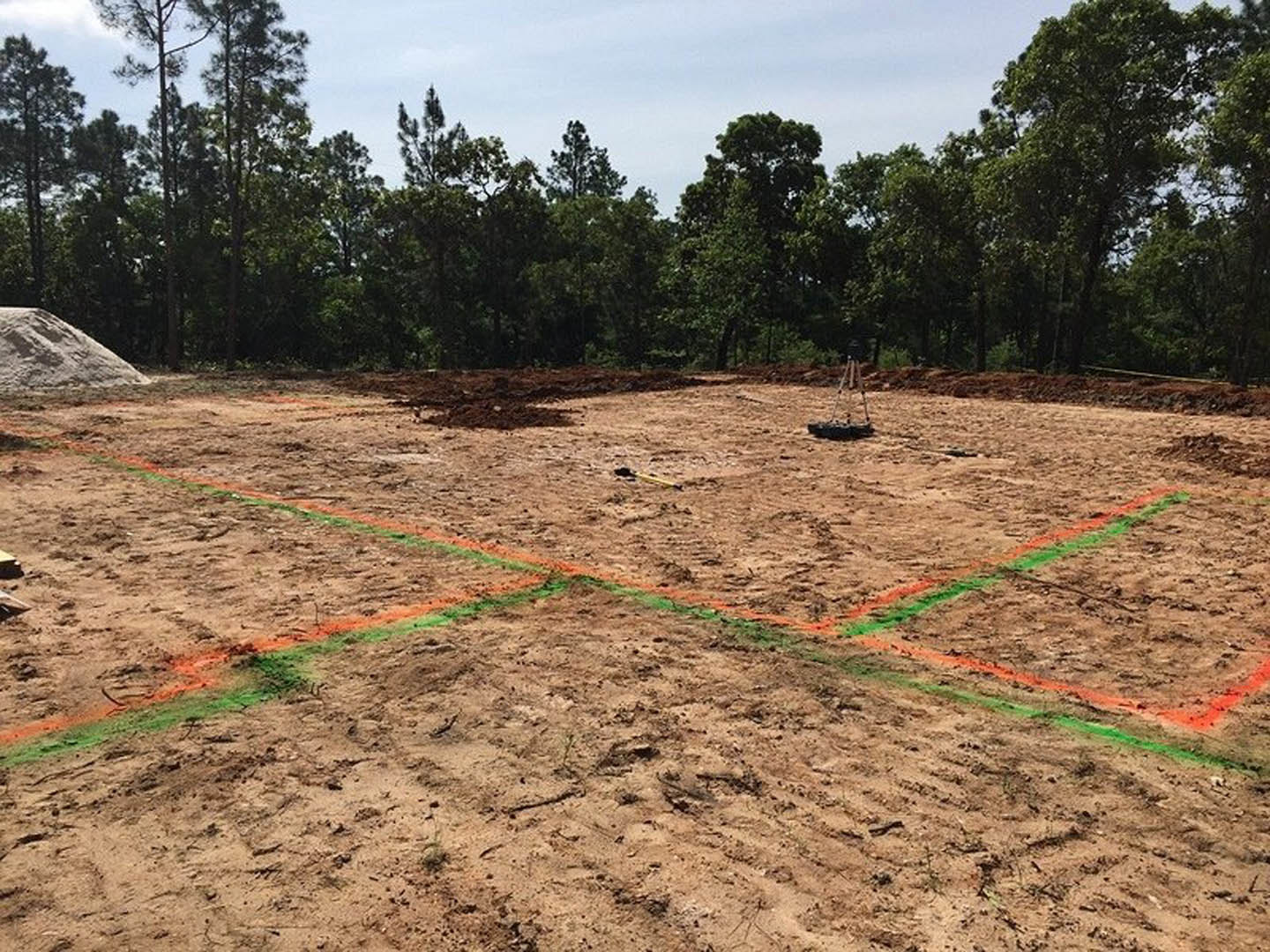 Dirt field with green and orange survey lines, tripod and land survey equipment in foreground, group of trees and large rock in background under open sky