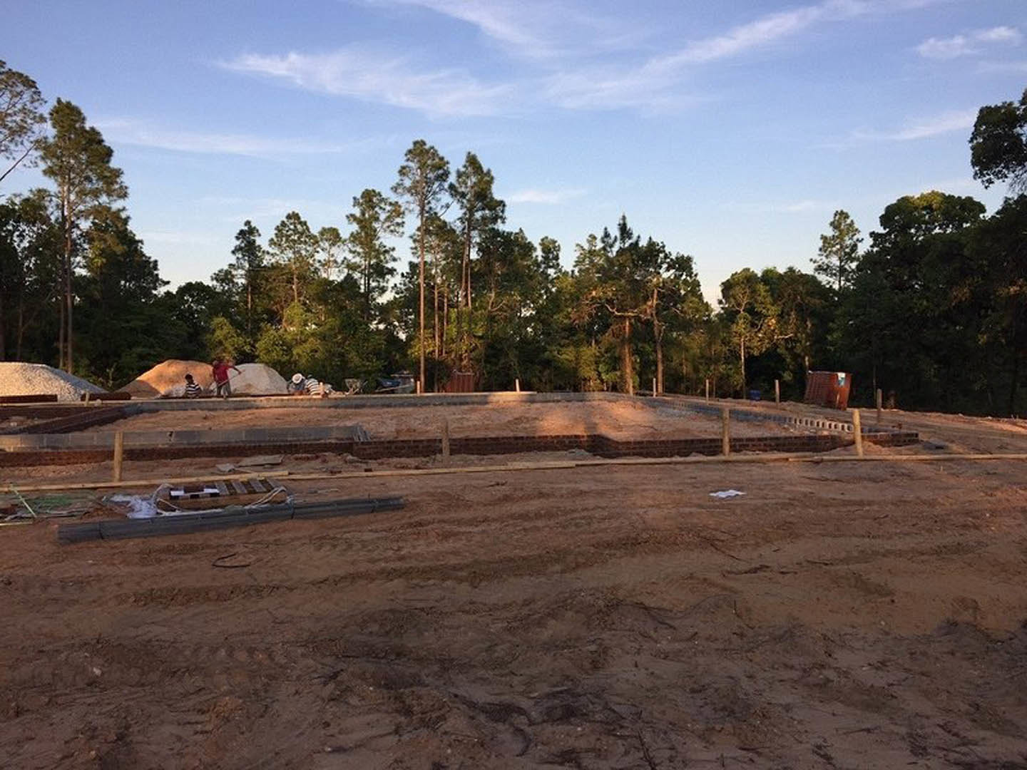 Dirt construction site bordered by a brick wall, surrounded by green trees under a blue sky with scattered clouds
