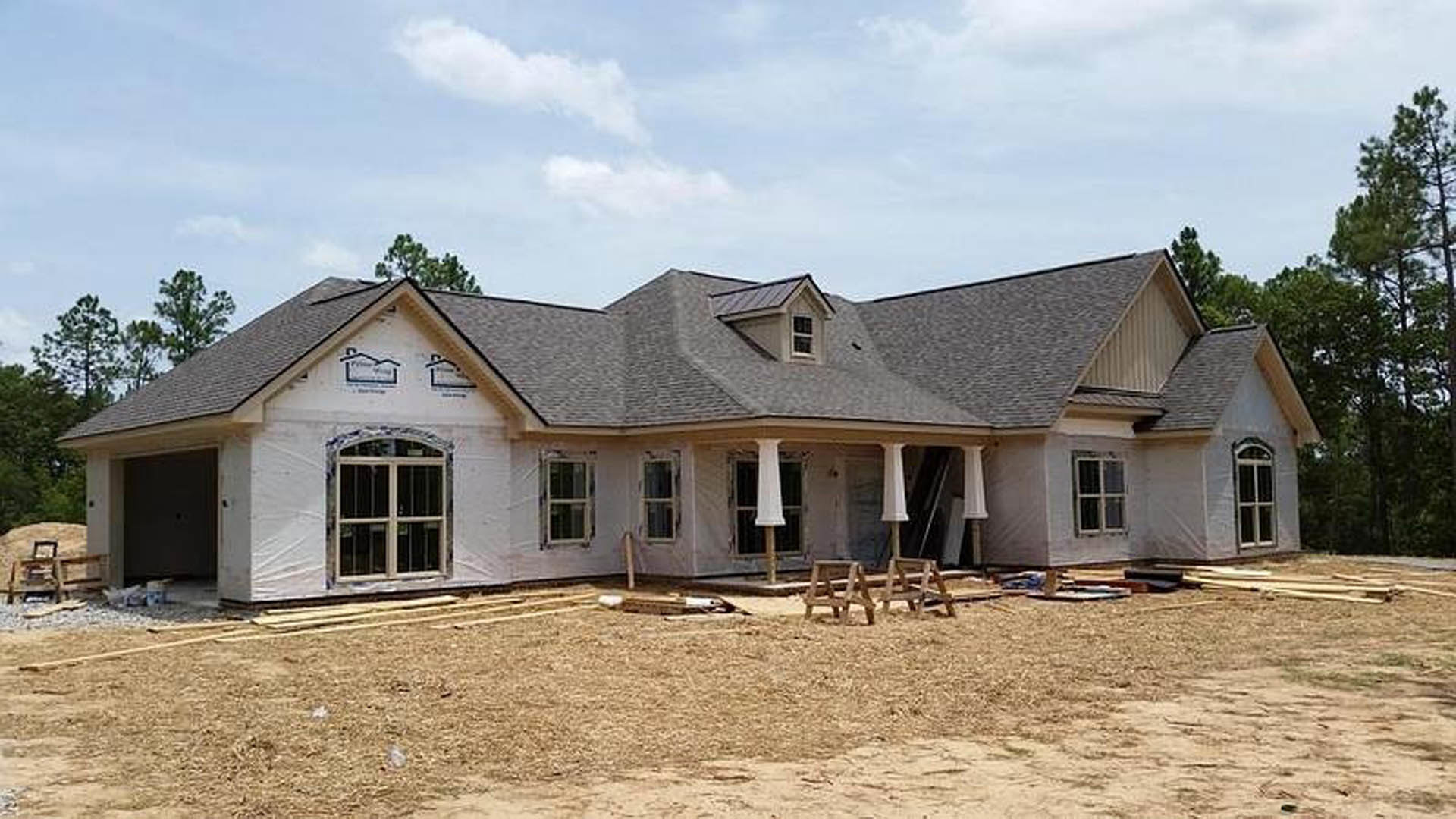 Partially built house with exposed framing and roof, dirt field in foreground, Robert Frost Farm and trees in background, blue sky with scattered clouds overhead