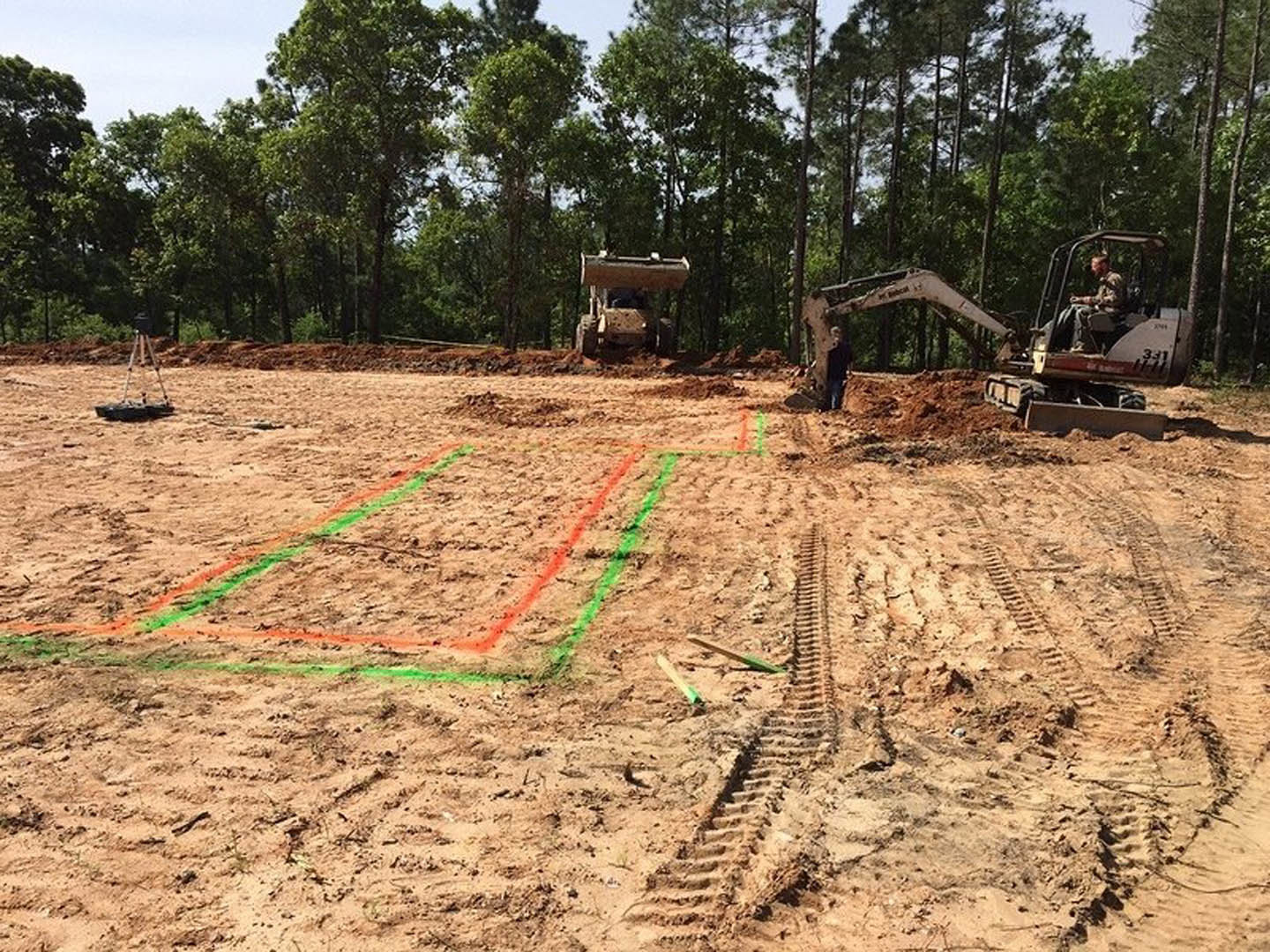 Construction site with muddy soil, tire tracks, and a tractor carrying a large log; trees and plants border the graded area under a cloudy sky
