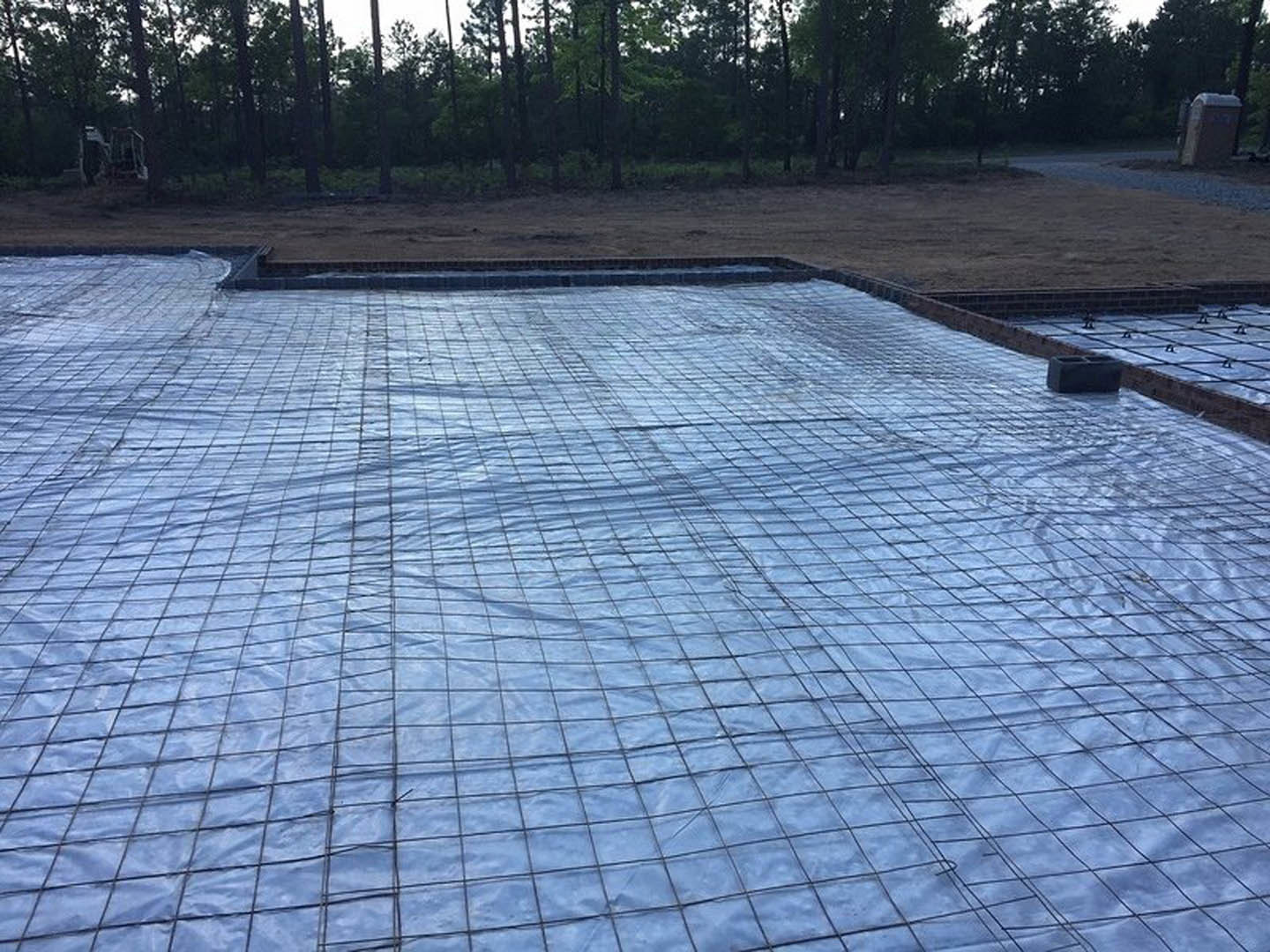 Black plastic tarp covering poured concrete foundation slab, surrounded by flagstone and trees in outdoor residential setting.