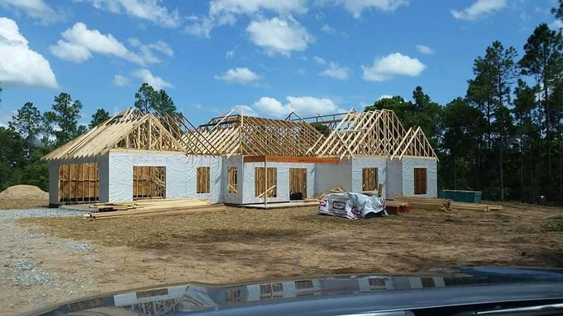 Wood-framed house under construction with exposed beams, barred window, brown wooden door, and parked car beside unfinished exterior