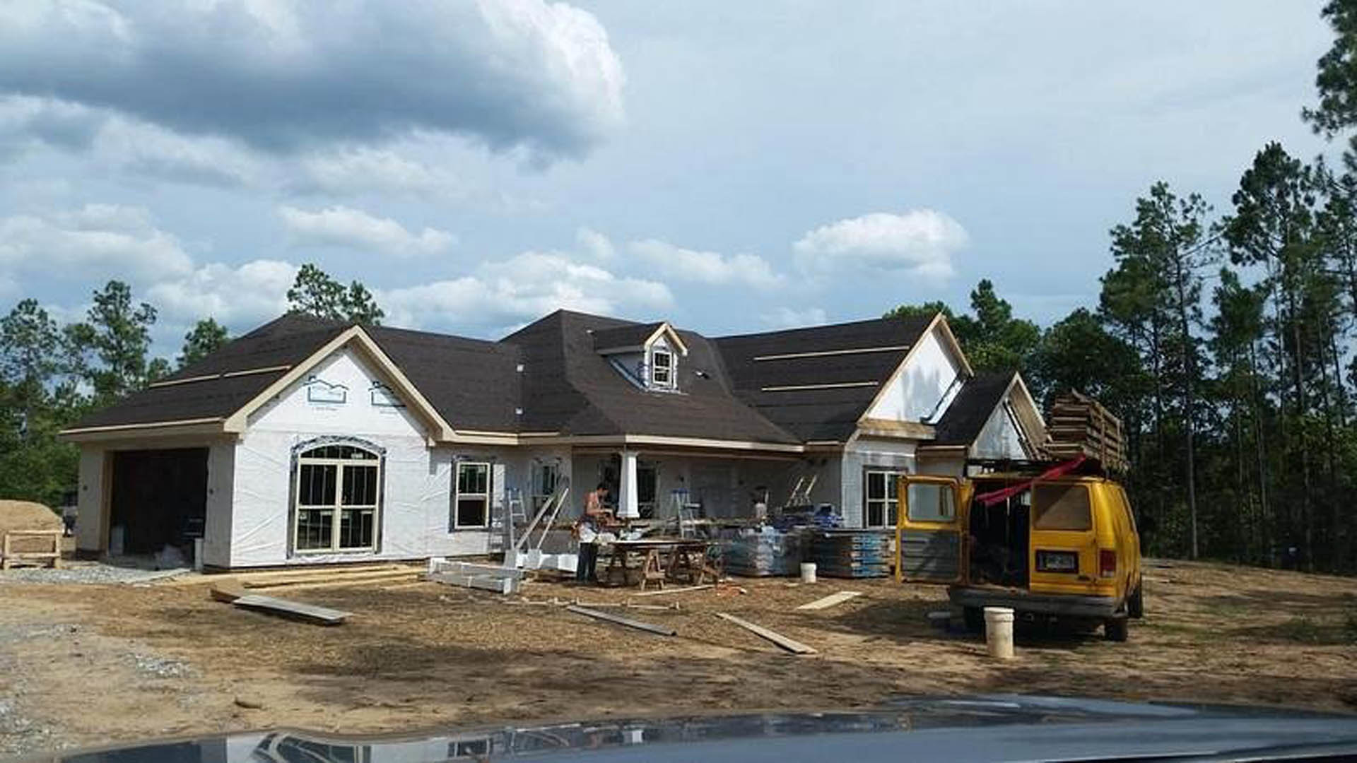 Partially built home with exposed framing, open window, yellow truck parked nearby, painter working on exterior, pillar in foreground, blue sky with scattered clouds