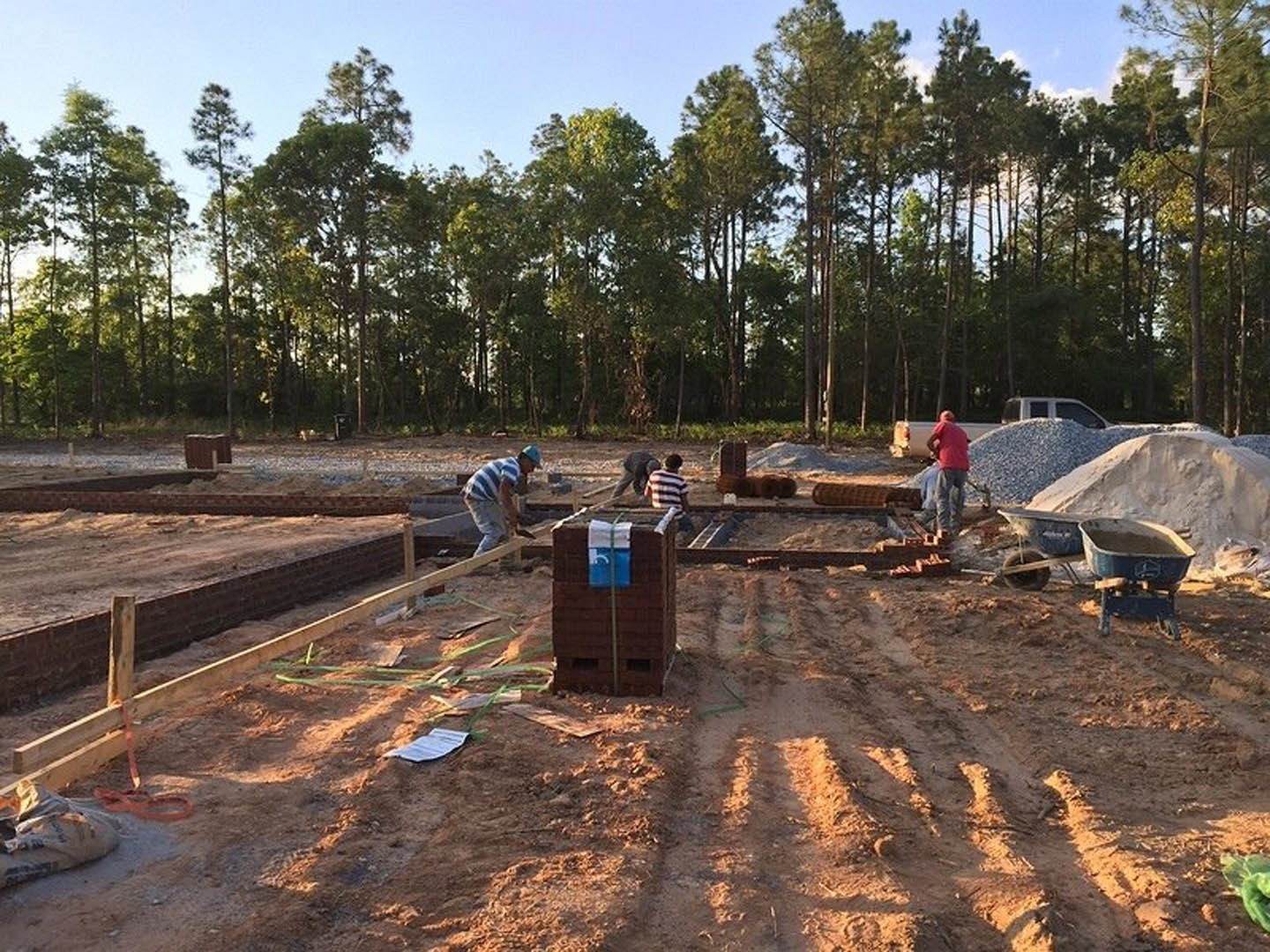 Construction workers building brick home foundation on muddy site, wheelbarrow with soil, blue safety helmets, trees in background, blue sign attached to structure