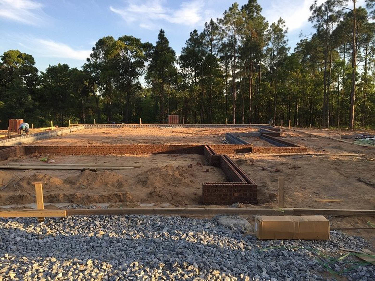 Partially built brick foundation surrounded by soil, scattered construction materials, and mature trees in the background under a cloudy sky
