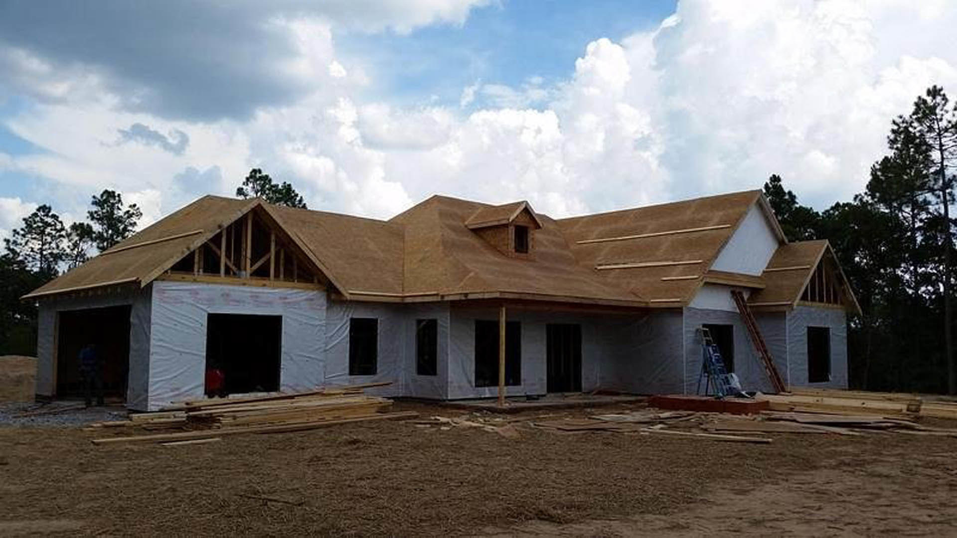 Partially built house with exposed framing, roof sheathing, and a ladder leaning against the exterior; dirt foreground, blue sky with scattered clouds