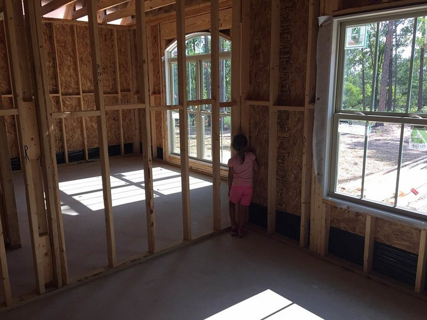 Girl in pink pants standing on wood floor, facing a white wall in a sunlit room with large window and tree views outside