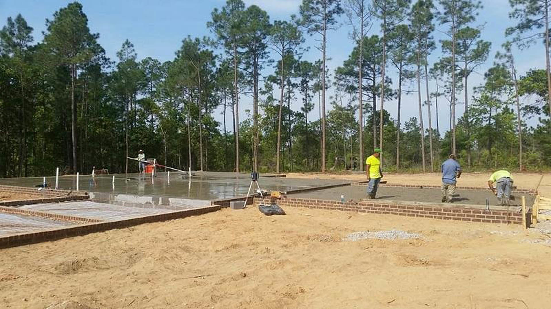 Partially built home foundation surrounded by soil and trees, several workers in yellow shirts moving and working on site, clear sky overhead